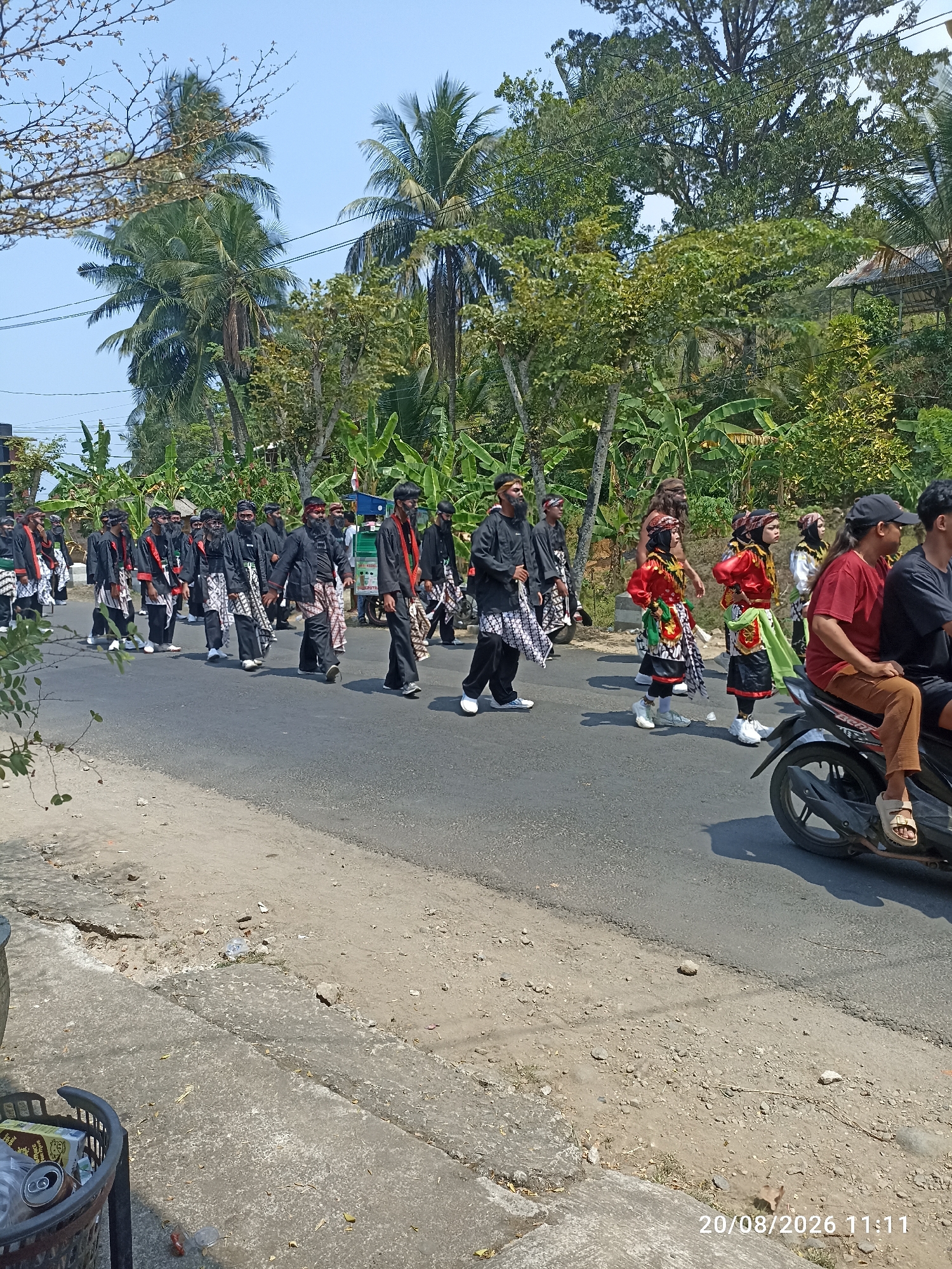 The image depicts a street procession in Munjungan, Indonesia, on August 20, 2026, at 11:11 AM. A group of individuals dressed in traditional black attire with patterned sarongs are walking down a paved road lined with lush green vegetation, including palm trees and banana plants. Some participants are wearing red sashes. To the right of the procession, a motorcycle carrying two individuals is visible, one wearing a red shirt and the other a black shirt. A woman in a red shirt is seated on the motorcycle, and a man wearing a black shirt is behind her. Further down the road, a cart is being pushed. The scene appears to be a cultural or religious event, given the traditional clothing. The sky is clear and blue. A black trash bin with some recyclables is on the left side of the image.