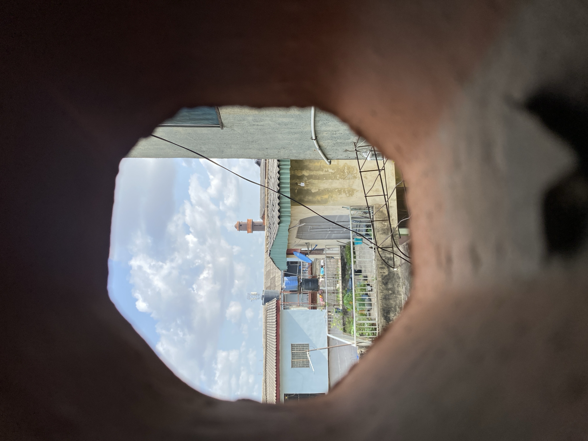 The image shows a view of a cityscape, possibly in Lagos, Nigeria, framed by an irregular, roughly circular opening in what appears to be a concrete or plaster structure. The perspective is slightly tilted. Within the frame, the view reveals buildings of varying heights and states of repair, typical of urban environments. Some buildings have corrugated metal roofs, while others appear to be concrete structures. Laundry is hanging on a clothesline between buildings. A minaret is visible in the distance. Power lines crisscross the scene. The sky is partly cloudy.