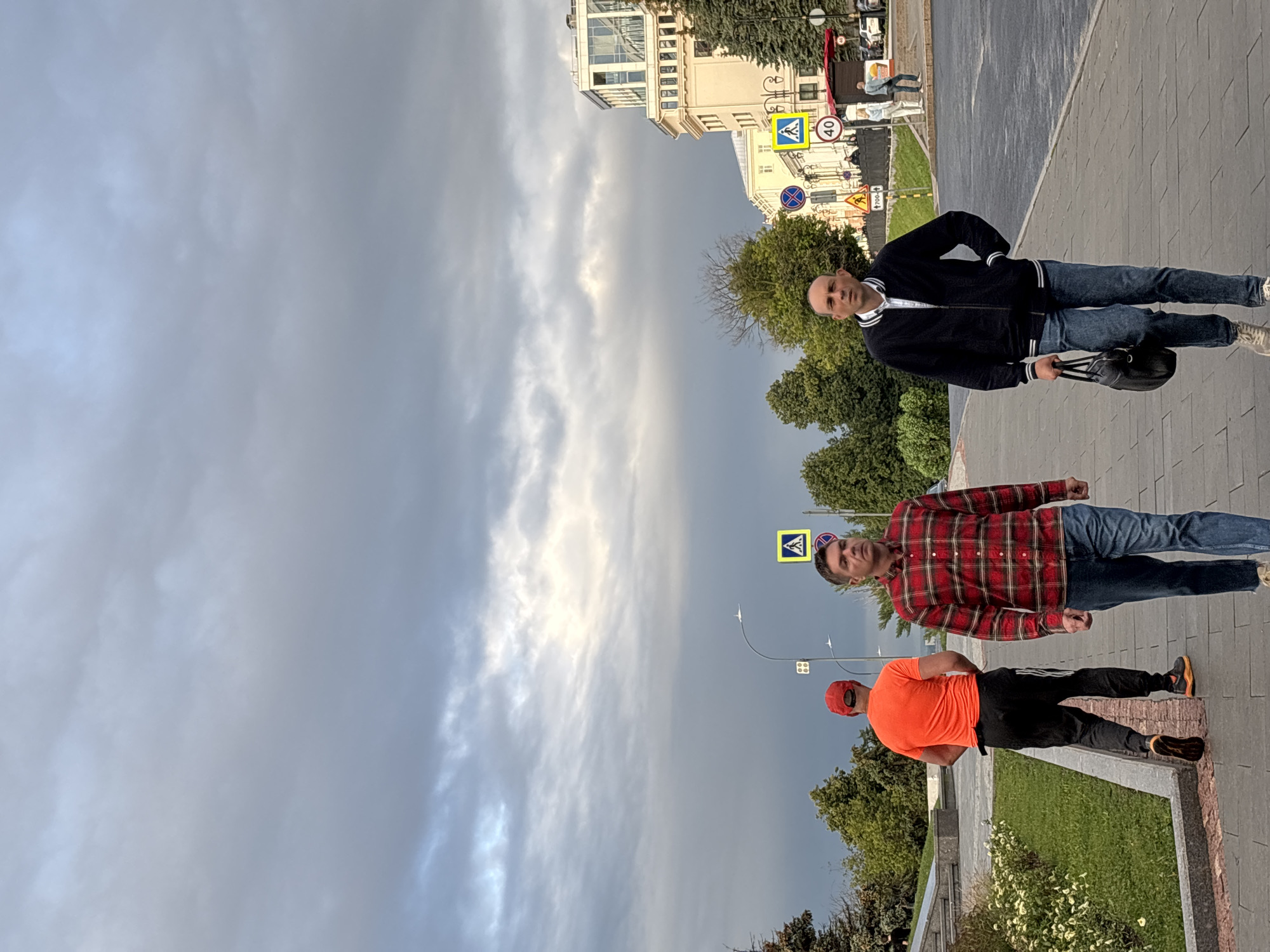 The image captures a street scene in Nizhny Novgorod, Russia, under an overcast sky with dramatic cloud formations. In the foreground, three men are walking on a paved sidewalk. The man on the far right wears a black bomber jacket with white trim over a white collared shirt and blue jeans, carrying a black leather bag in his right hand. Next to him, a man in a red and black plaid long-sleeved shirt and blue jeans walks towards the camera. Further to the left, a man wearing an orange t-shirt and black pants with white stripes on the side, and a red cap, is seen from behind, walking away from the camera.
Behind the men, a paved road with a crosswalk and traffic signs is visible. Among the signs are a pedestrian crossing symbol and a speed limit sign indicating "40". Several trees line the sidewalk and the area beyond the road. In the background, a multi-story building with a modern facade, featuring white and glass elements, stands prominently. The overall atmosphere suggests a cool or temperate climate, possibly late afternoon or early evening. The perspective of the photograph is from a slightly low angle, looking up towards the sky and the building.
