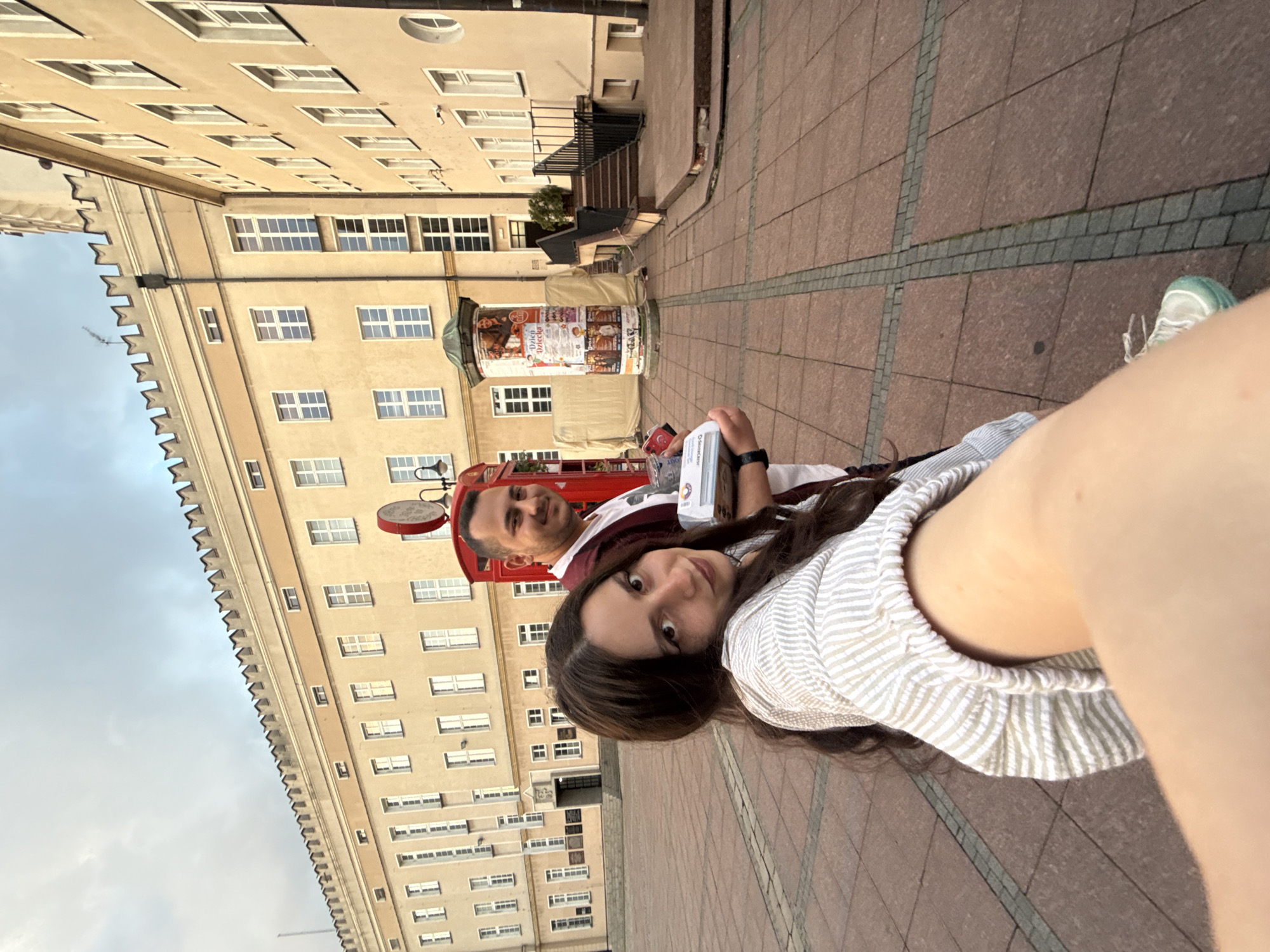 A selfie captures a young woman and man in an outdoor urban setting in Opole, Poland. The woman, in the foreground on the right, has long dark hair and wears a striped top. She looks directly at the camera with a subtle smile. Behind her, a man with short dark hair and a beard smiles faintly towards the camera, wearing a dark top over a lighter shirt. He is partially obscured by the woman and a red structure behind him, which might be part of a vehicle or a stylized phone booth, featuring a small white circular sign with illegible dark text.
They are standing on a paved ground composed of reddish-brown and gray square tiles arranged in a grid pattern. In the background, several multi-story buildings with numerous windows line the street. The building immediately behind the couple is light-colored, possibly beige or cream. To the left, a more ornate building with a distinctive crenellated roofline stands against a light, overcast sky.
A prominent cylindrical advertising column (kiosk) is visible behind the man, adorned with posters. One poster clearly displays the Polish word "Dzieci" (Children) and likely refers to "Dzień Dziecka" (Children's Day), along with other graphical elements and text that are not fully decipherable. The overall lighting suggests late afternoon or early evening, casting a soft glow on the scene. The atmosphere appears relaxed and casual.