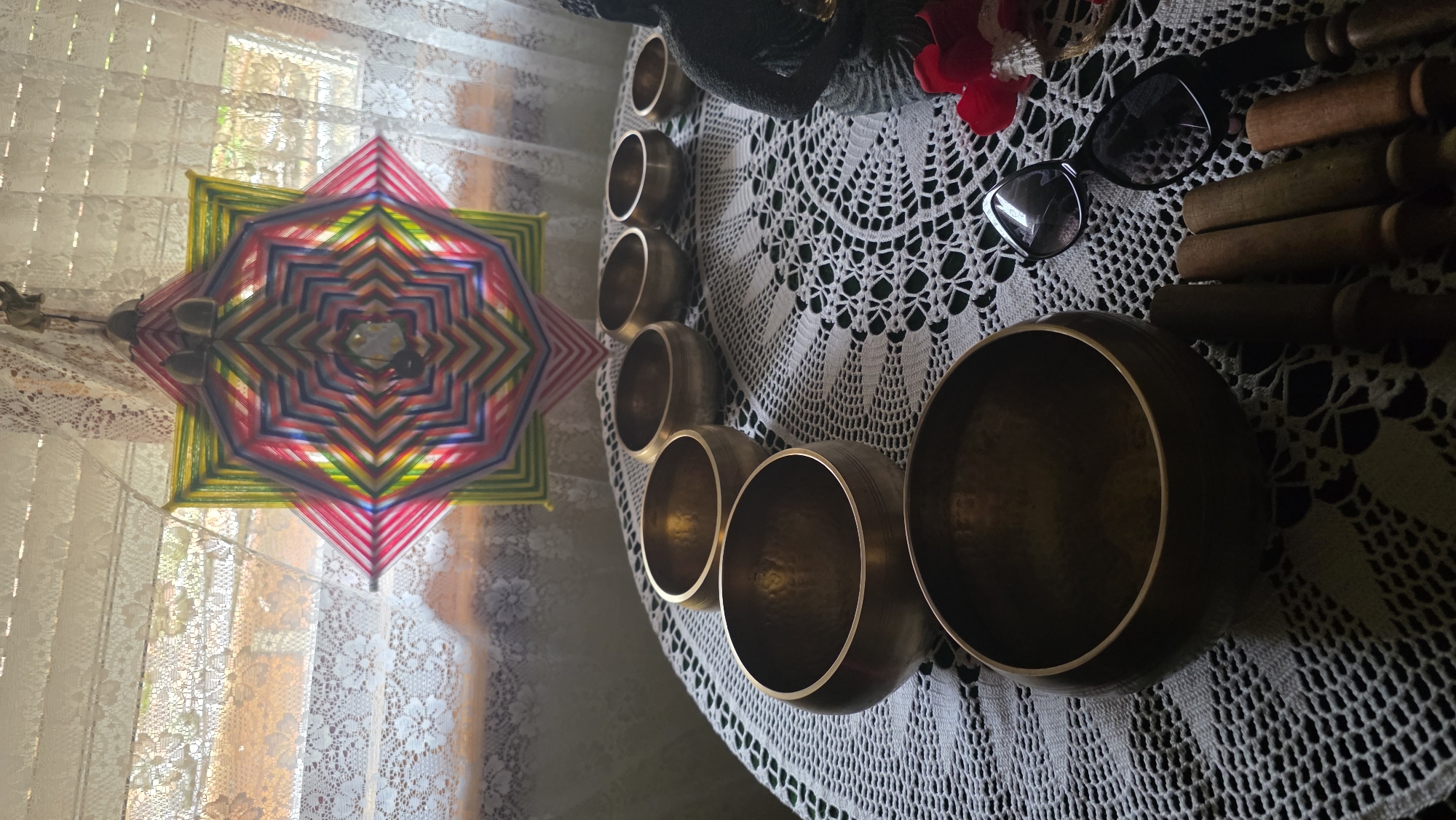 The image captures an indoor scene in Blagoevgrad, Bulgaria, characterized by objects commonly associated with mindfulness and spiritual practices. The photograph is taken from a slightly elevated perspective, looking down onto a surface covered by an intricate white crocheted tablecloth or doily.
Dominating the right side of the frame is an array of brass singing bowls, varying in size, arranged on the white lace. Their metallic surfaces reflect light, suggesting a well-lit environment. Alongside these bowls are several wooden mallets or strikers, tools typically used to play the singing bowls. Also present on the tablecloth are a pair of dark-framed sunglasses and a small cluster of artificial red flowers, with a dark, indistinct object partially visible behind them.
To the left, against the backdrop of a window, hangs a vibrant, multi-colored geometric yarn art piece, resembling a "God's Eye" or a mandala. This symmetrical artwork displays a spectrum of colors including pink, red, yellow, green, and blue. The window itself is adorned with light-colored lace curtains, through which bright natural sunlight streams, indicating a daytime setting and contributing to a peaceful, warm atmosphere.
No direct activity is depicted, but the arrangement of items strongly suggests a space prepared for meditation, sound healing, or a similar contemplative practice. There is no visible text in the image.