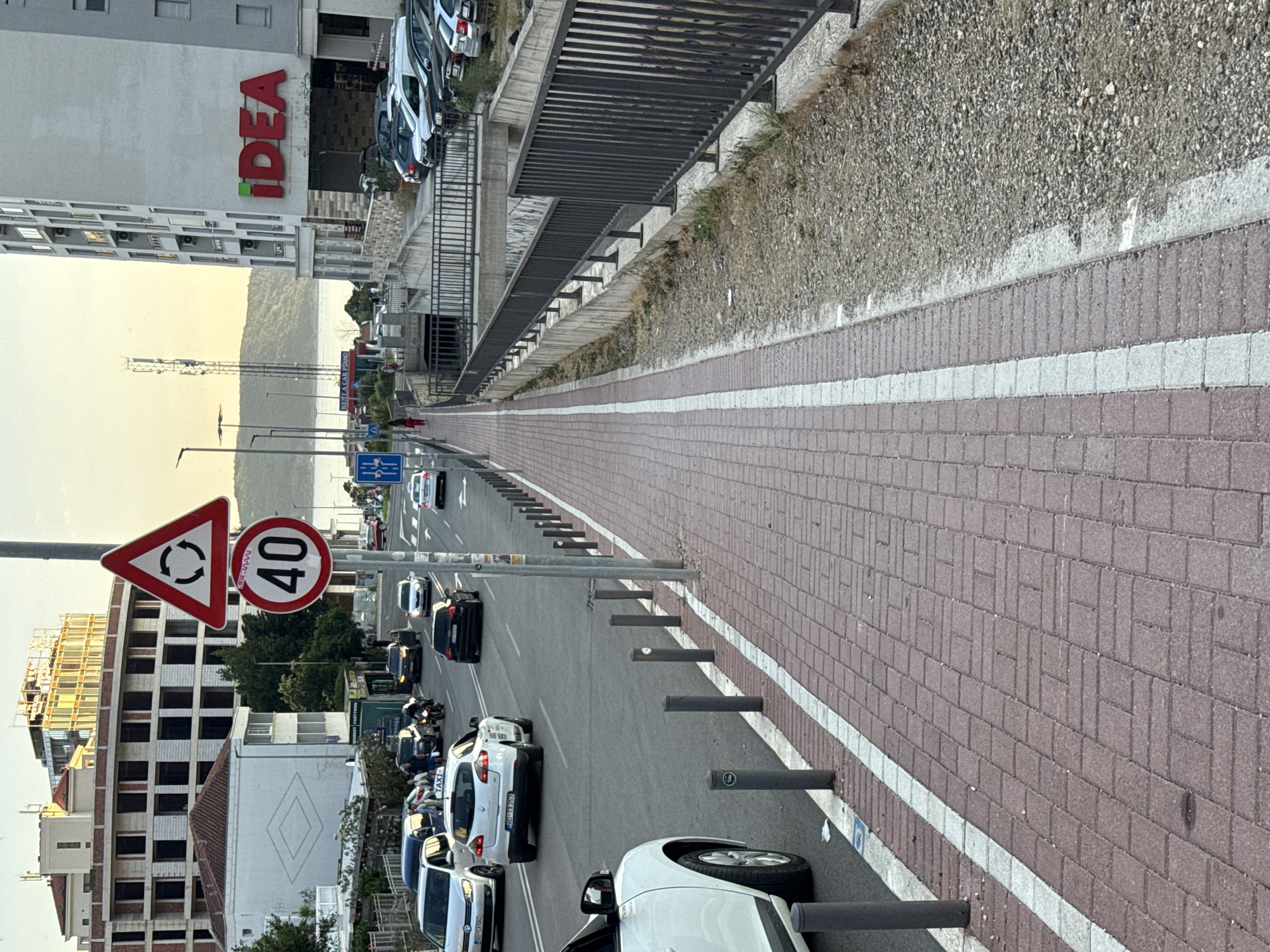 The image depicts a street scene in Boreti, Montenegro, with a focus on a pedestrian walkway and the adjacent road. The walkway is paved with reddish-brown bricks and has a white stripe running along its edge, separating it from the road. A metal fence runs alongside the walkway, bordering a lower, gravel-covered area.
On the road, several cars are visible, including a white hatchback with a "TAXI" sign on its roof, a black sedan, and other vehicles of various colors. A motorcycle is also present in the traffic. Traffic signs are prominent: a triangular sign with rotating arrows indicates a roundabout, and a circular sign displays a speed limit of 40. A person in a red outfit is walking on the pedestrian walkway, heading away from the camera.
In the background, there are buildings, including a large building with the prominent "IDEA" logo. The sky is visible, with a hint of the sea in the distance and a hilly landscape beyond the water. The overall scene suggests a moderately busy urban environment, possibly during daylight hours.