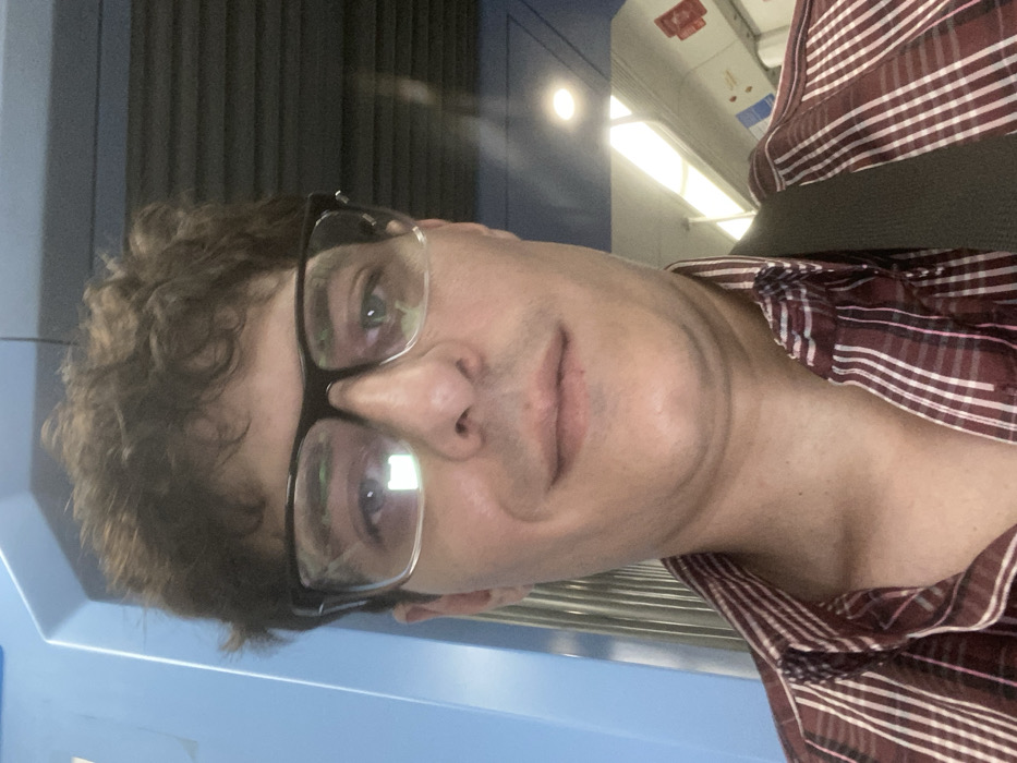 A male individual is depicted in a close-up, mid-shot, looking directly forward. He has medium-toned skin, dark, curly hair, and wears black-framed glasses. His expression is neutral with a slight upward turn at the corners of his mouth. He is dressed in a collared shirt featuring maroon/burgundy and white plaid stripes, and a dark strap, possibly from a bag, is visible over his right shoulder. The background indicates an interior setting, identified as a public transport vehicle, consistent with a train or subway car, located in Llavallol, Argentina. To the left, a light blue vertical panel is visible. To the right, the vehicle's interior features a white ceiling with both circular and elongated fluorescent-style light fixtures, off-white wall panels, and red and white rectangular signs or markings. Portions of horizontal metal bars, likely handrails, are also visible.
