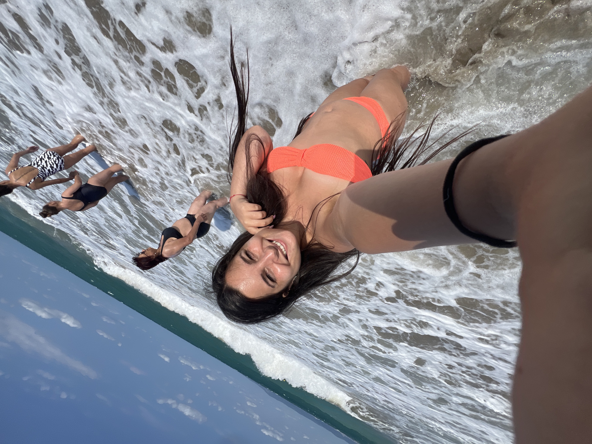 In Pomorie, Bulgaria, four women are depicted in the shallow waters of the Black Sea on a sunny day. The perspective is from a selfie taken by the woman in the foreground, who is smiling and wearing an orange bikini. Her dark hair is wet and extends outwards. Behind her, three other women are visible. One woman in a black bikini is to her left, facing away. Further back, another woman in a black one-piece swimsuit and a fourth woman in a black and white patterned swimsuit are also present. The sea is slightly choppy with waves and foam washing over the women's legs. The sky is blue with scattered clouds, and a faint outline of land is visible in the distance.