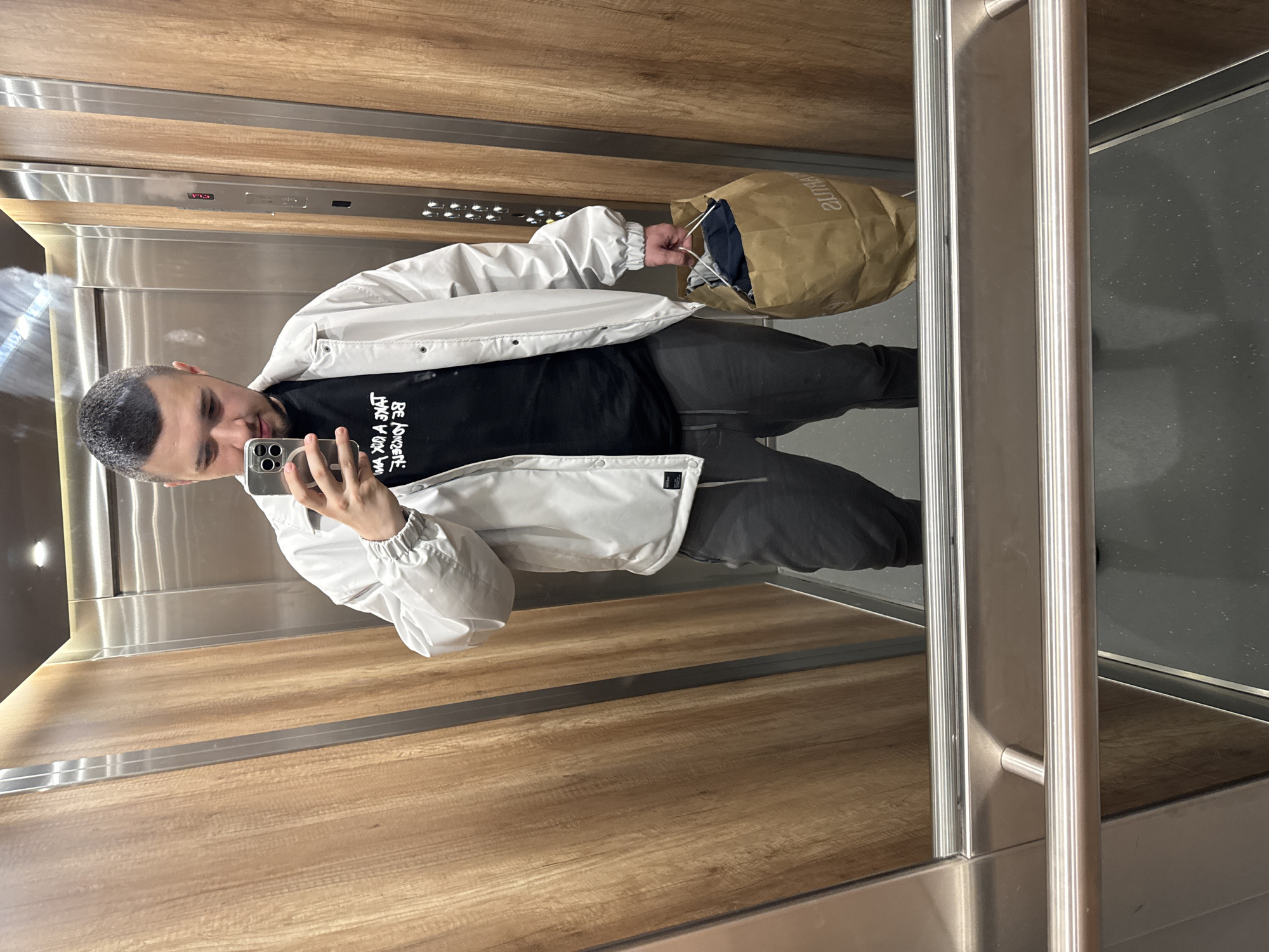 A man is captured in a mirror selfie inside an elevator, likely in Sofia, Bulgaria. The image is rotated 90 degrees clockwise.
**Subjects:** The primary subject is a man with short, dark hair and neatly trimmed facial hair. He is holding a silver smartphone with a multi-lens camera, using it to take a selfie in the elevator's mirrored wall. His expression is neutral, focused on the phone.
**Objects:** He is wearing a light-colored (possibly white or light grey) jacket with elasticized cuffs over a black t-shirt. Dark grey or black trousers complete his attire. In his left hand, he holds a brown paper shopping bag, which has some items visible inside and a partially mirrored logo or text that appears to say "SOUTHAN" or similar. The smartphone is prominently visible in his right hand.
**Scene and Setting:** The scene is set inside a modern elevator. The walls are a mix of light wood-paneled sections and brushed metal panels. The floor is a dark grey, possibly speckled or textured. A silver handrail runs vertically along one side. An elevator control panel is visible with a digital display showing "14" or "143", indicating the current floor, along with several unidentifiable buttons.
**Activity:** The man is taking a mirror selfie, typical of checking one's appearance or capturing an outfit of the day (OOTD).
**Notable Details:** The lighting is artificial, consistent with an indoor setting like an elevator. The reflection in the mirror distorts some elements, particularly the text.
**Visible Text:**
* On the man's black t-shirt, mirrored English text is partially visible, appearing to read "BE LOVEDIT LIVE YOUR LIFE" or a similar motivational phrase.
* On the paper shopping bag, a mirrored brand name or text is visible, possibly "SOUTHAN".
* The elevator control panel shows the number "14" or "143" on a digital display.