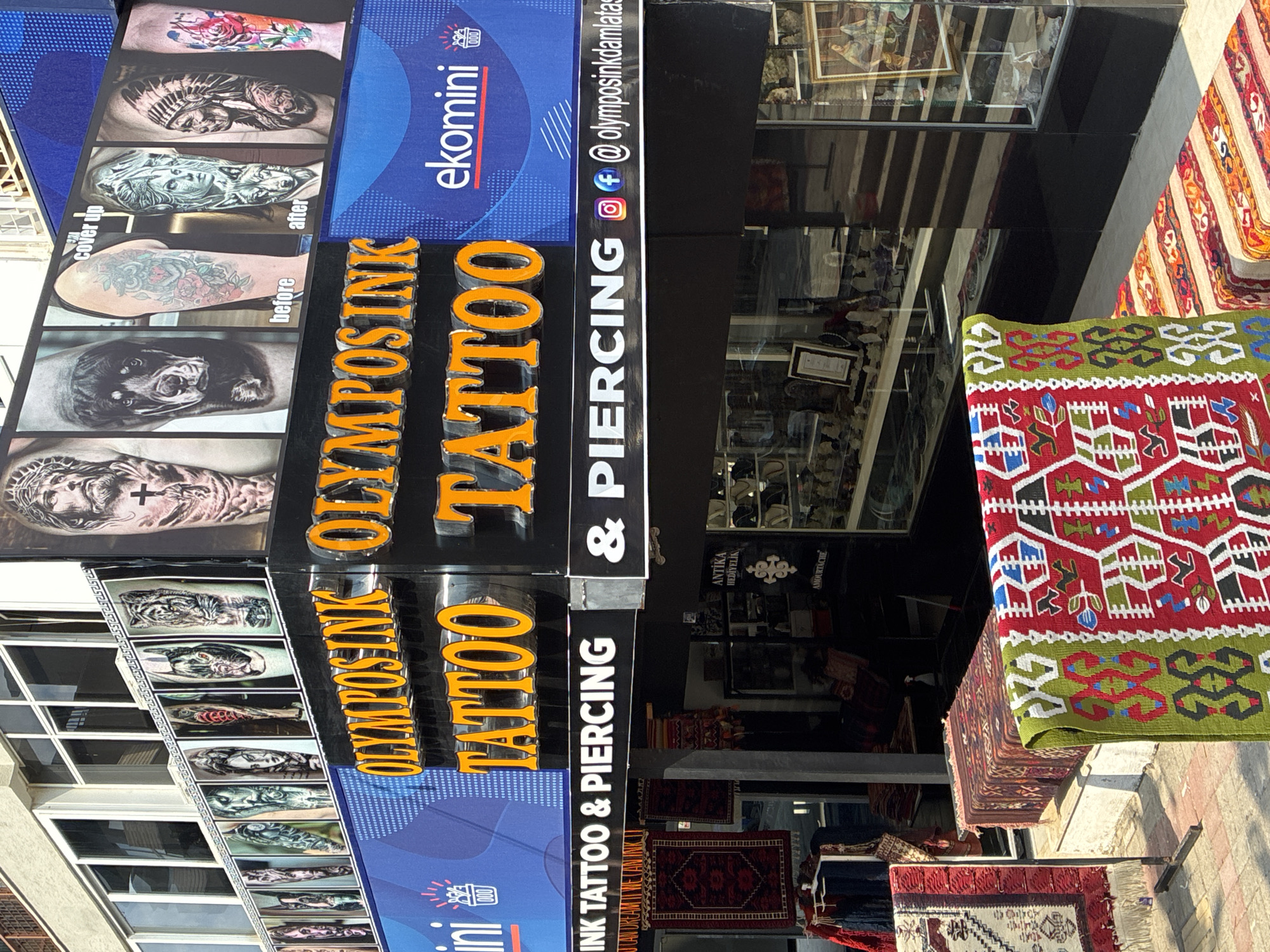 The image shows the storefront of "Olympus Ink Tattoo & Piercing" located in Alanya, Turkey. The facade features large, illuminated signage with the business name in orange, three-dimensional lettering. Above the signage, a display showcases various tattoo designs, including portraits of Jesus, a dog, a tiger, a woman, and an Indigenous American. Below the main sign, the words "INK TATTOO & PIERCING" are displayed in white lettering, accompanied by social media icons for Instagram and Facebook, and the business's social media handle. The storefront itself appears to be a shop, with racks of colorful textiles and rugs displayed outside. Through the windows, shelves stocked with various items, possibly jewelry or souvenirs, are visible. The overall scene suggests a bustling commercial area in a Turkish city, likely a tourist destination given the presence of rugs and the nature of the business.