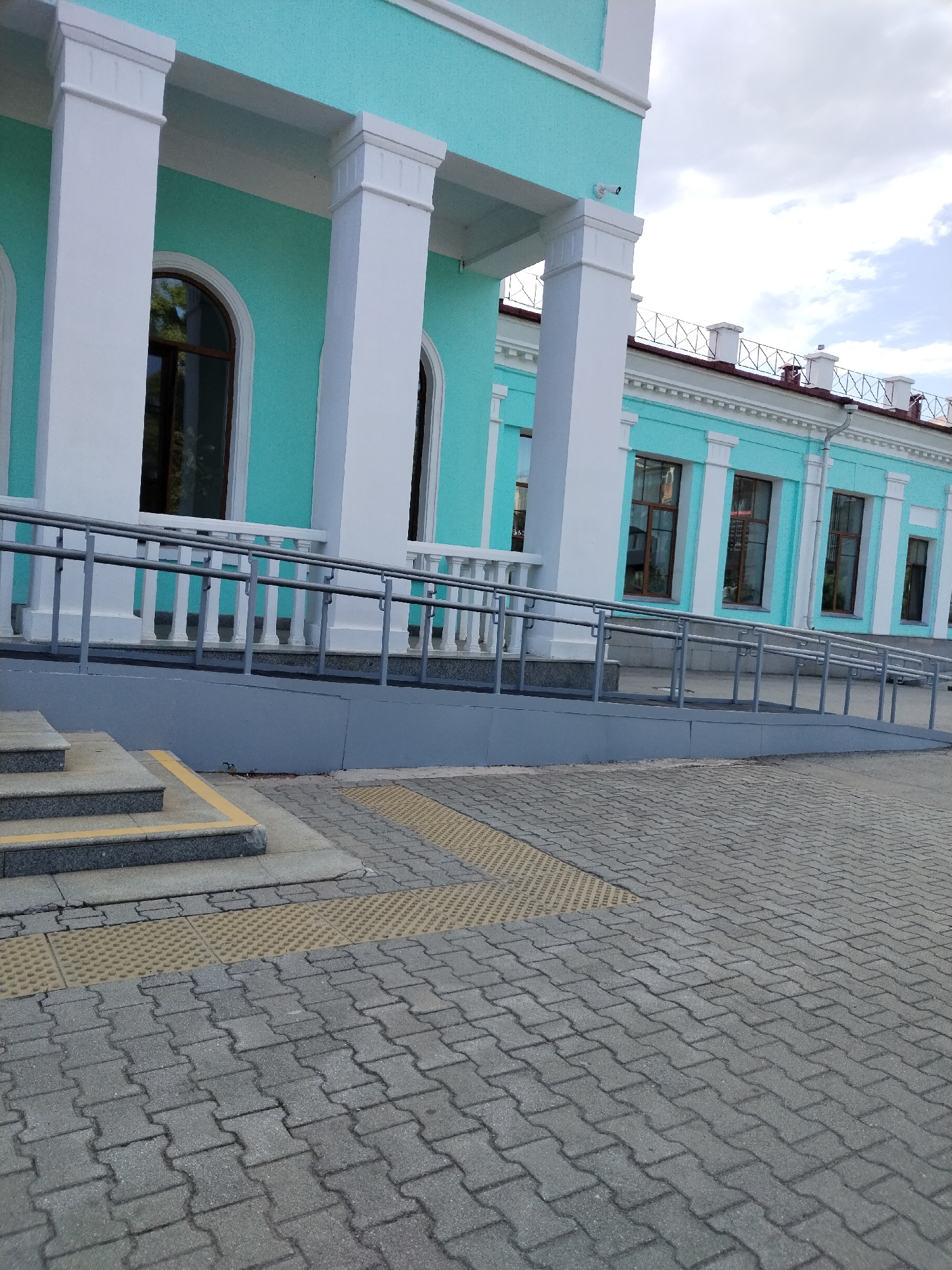 The image depicts the exterior of a building with a turquoise facade and white columns. In the foreground, there are stone steps leading up to a ramp with metal railings, situated next to the building. The ground is paved with interlocking gray stones and features tactile paving for visually impaired individuals. A security camera is visible mounted on the building near one of the columns. The sky is cloudy, suggesting overcast weather. The scene appears to be in an urban environment, possibly a public building such as a train station or municipal office. There are no people visible in the image.