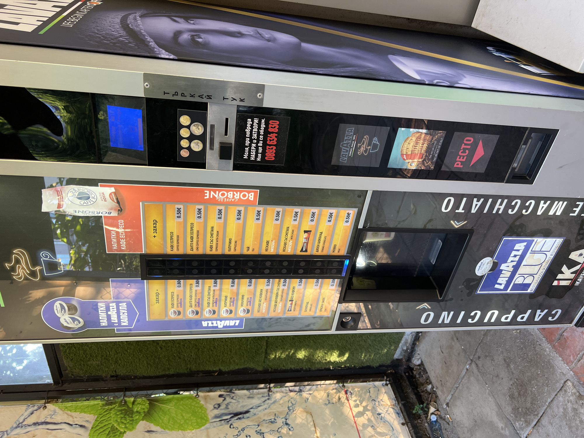 This is a photograph of a coffee vending machine located on a sidewalk in Burgas, Bulgaria. The machine offers a variety of hot beverages, including espresso, cappuccino, tea, and hot chocolate. The prices are displayed in Euros. A sign on the machine provides a phone number for customer service. The sidewalk is paved with concrete and brick, and there is some greenery around the machine. The background is blurred, but it appears to be a street scene with buildings. The weather is sunny.