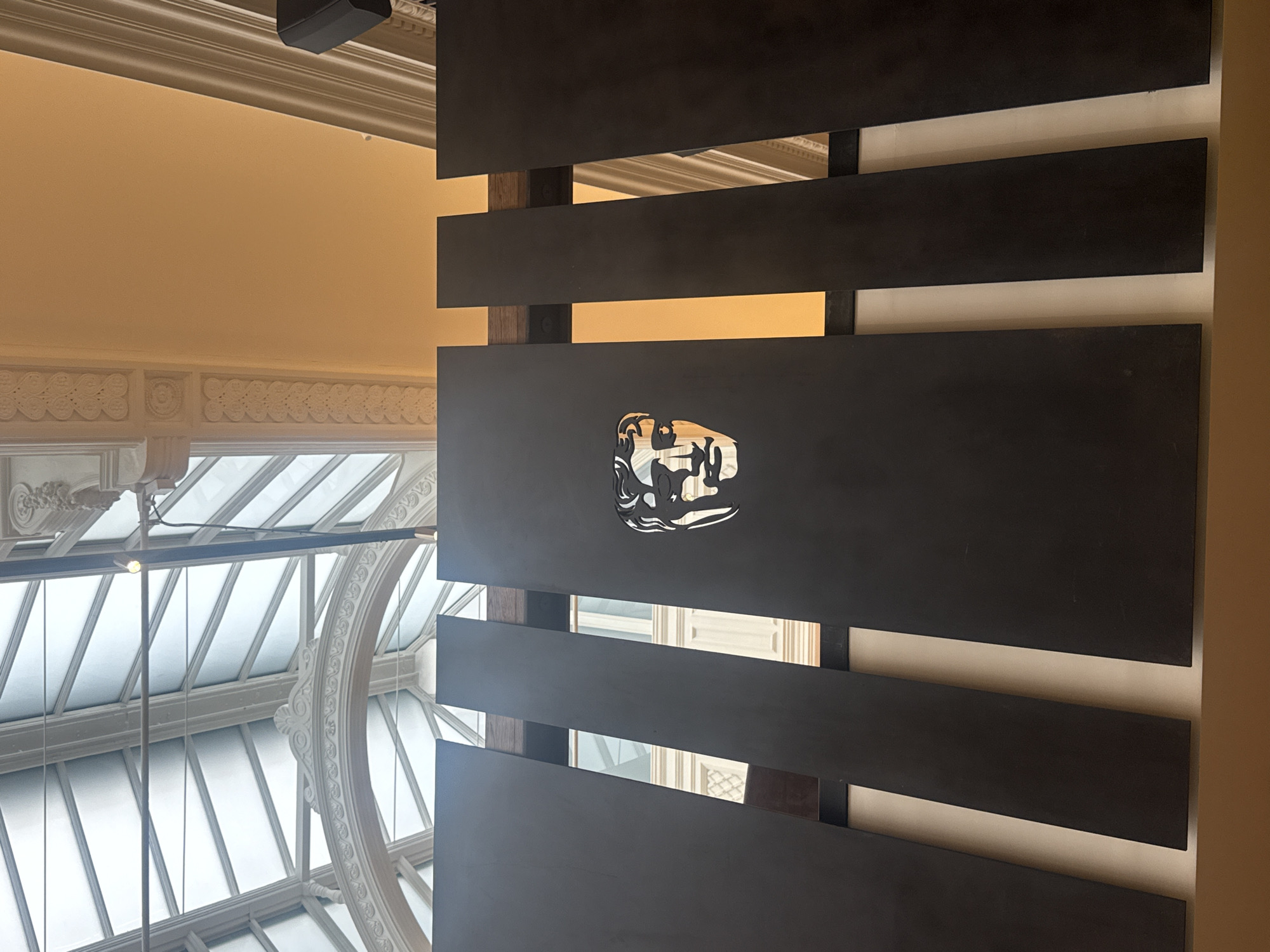 The image presents an upward view of a grand interior space, combining historical architecture with modern design elements. A prominent feature is a modern partition composed of multiple dark, horizontal rectangular panels that are spaced apart, revealing a lighter-colored wall or background behind them. Centered on one of these dark panels is a distinctive, highly reflective metallic logo or symbol. This emblem appears to be a stylized head or mask, possibly animal-like or mythological, with intricate lines and an abstract design that reflects the surrounding light.
To the left, a magnificent arched glass skylight or ceiling dominates the view, featuring elaborate white architectural moldings, including a large decorative circular arch and detailed ornamental patterns. A warm, light beige wall or ceiling is visible above the partition, also adorned with classic decorative cornices. A black speaker is partially visible in the upper-left corner of the frame. The bright light streaming through the skylight indicates it is daytime, illuminating the scene and creating strong reflections on the metallic logo. The architectural details suggest a significant historical building, possibly in London, United Kingdom, as provided by the context. No people or specific activities are depicted.