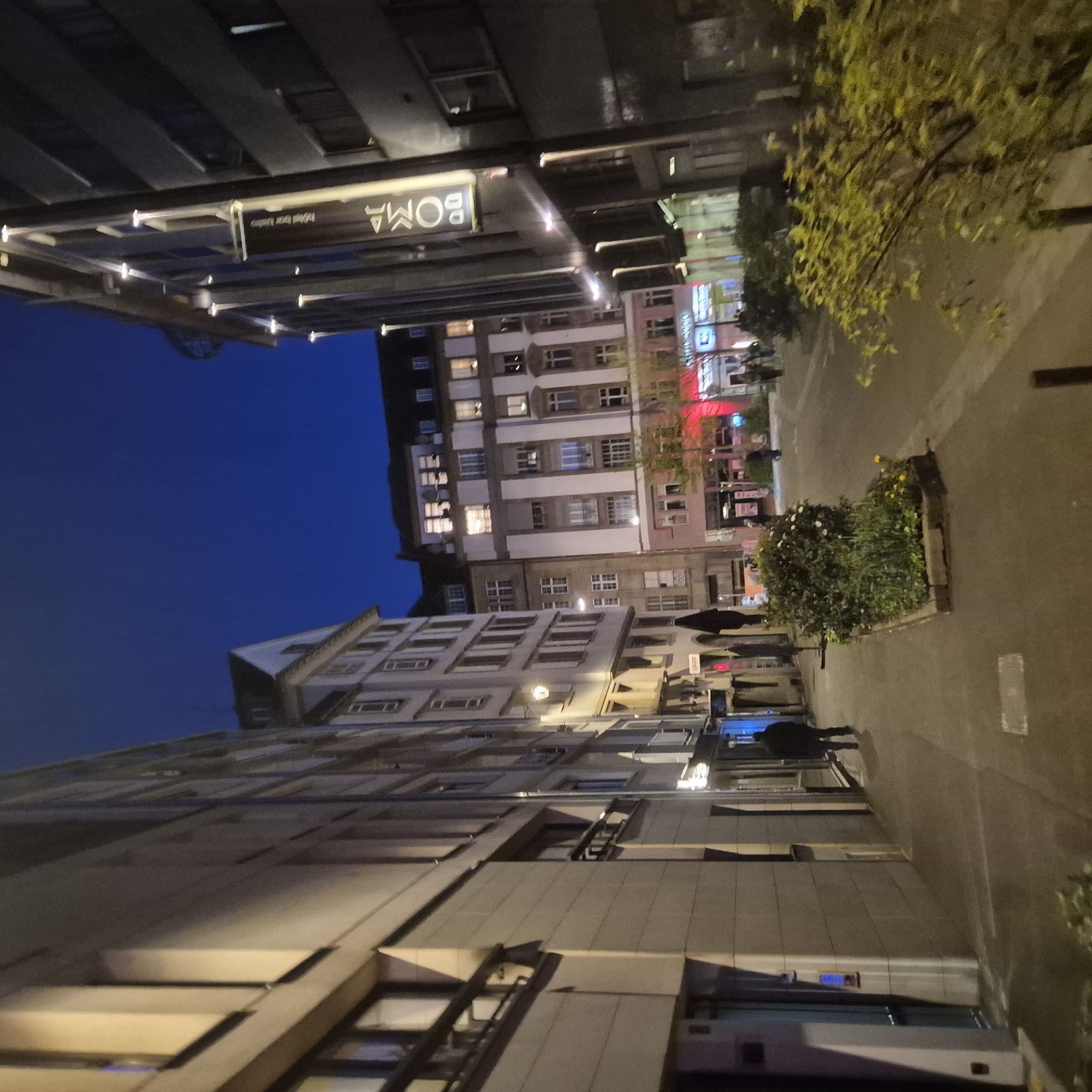 The image shows an urban streetscape in Strasbourg, France at night. The sky is dark blue and the street is lit by streetlights and building lights. Buildings line both sides of the street. The "BOMA" sign (likely the Hotel BOMA) is visible at the top-left of the frame. Pedestrians are walking on the pavement, with some holding umbrellas. Flower beds are visible along the street. The perspective is slightly tilted.