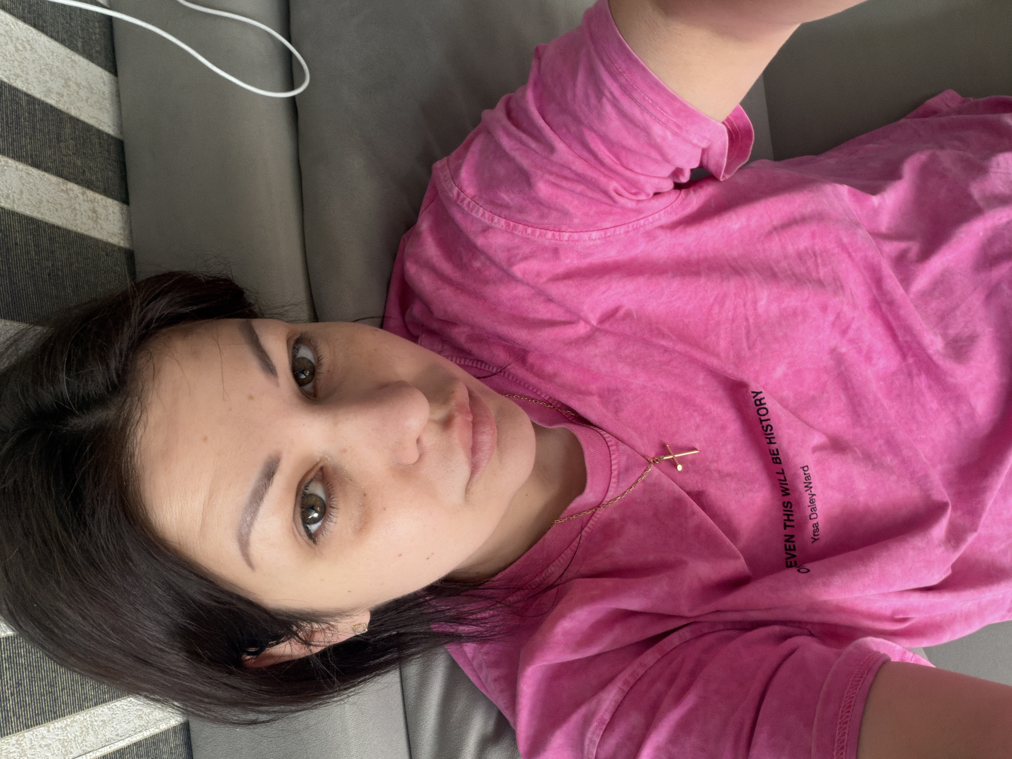 A young woman with dark hair is lying down and looking at the camera. She is wearing a pink t-shirt with text on it, a gold necklace with a cross pendant, and a small gold earring. Her eyes are brown, and she has dark circles under them. The background is a textured surface that appears to be a sofa or bed, with some striped material visible above her head. A white cord is also visible in the background. There is no indication that this image was taken in Sochi, Russia.