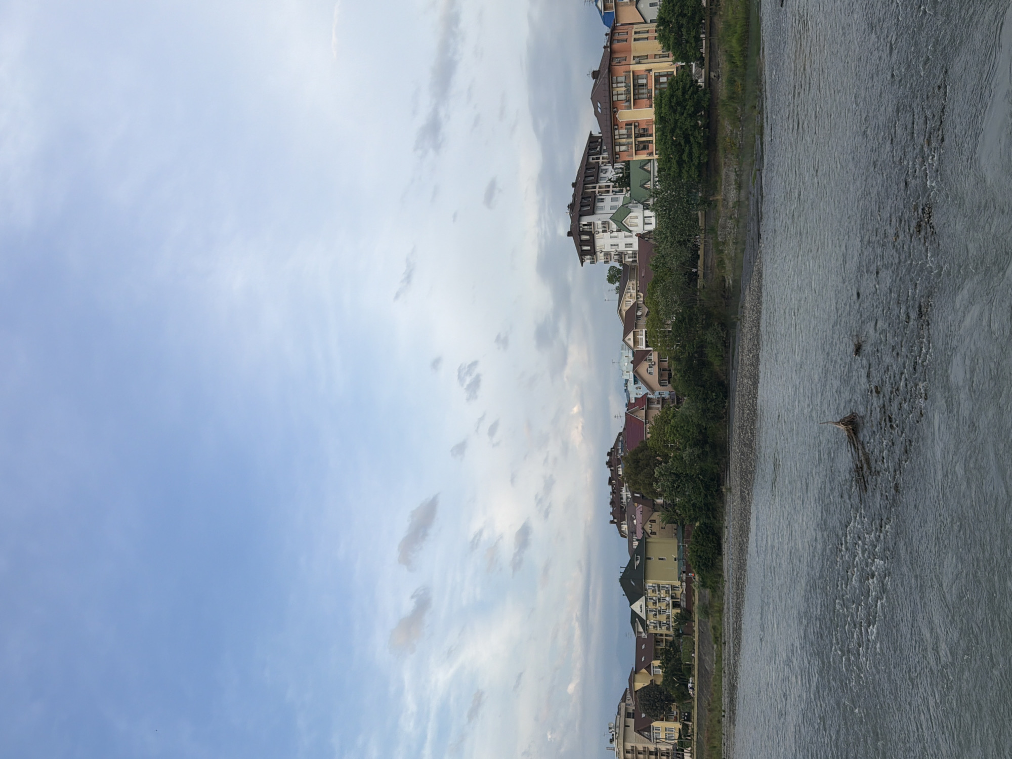 The image depicts a wide, fast-flowing river in the foreground, with a treelined embankment and a row of multi-story buildings beyond. The sky is a light blue with scattered white clouds. The buildings appear to be residential or hotel accommodations, with varied architectural styles and colors. The embankment is covered with gravel and vegetation. In the river, there are some submerged rocks and a piece of driftwood. The scene suggests a moderately populated urban area near a natural water source in Sochi, Russia, likely during daylight hours.