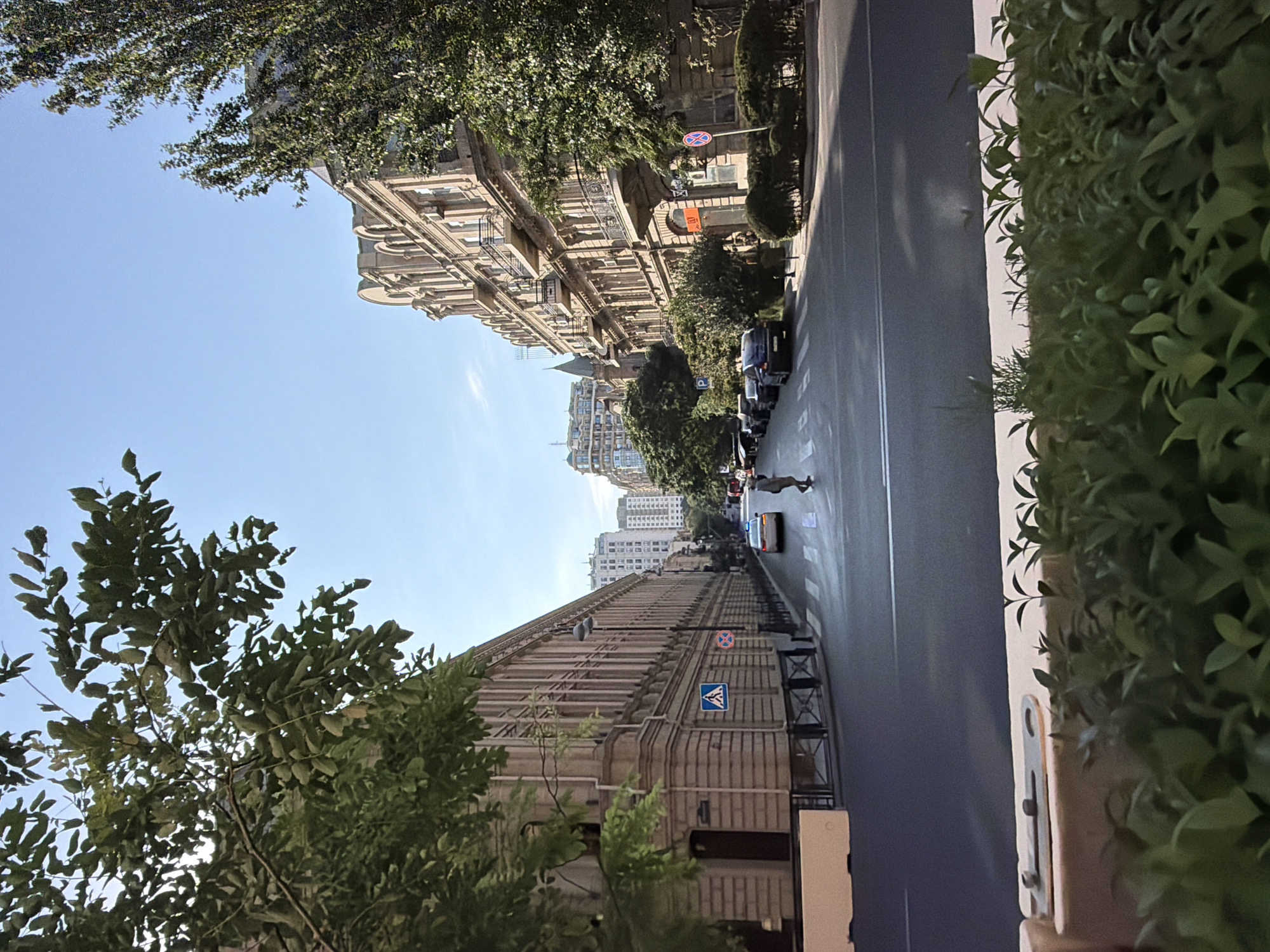 The image captures a view down a city street in Baku, Azerbaijan. Two multi-story buildings with ornate architecture line either side of the road. The building on the left is a lighter stone color and features many regularly spaced vertical lines, suggesting windows or architectural elements. The building on the right is a similar stone color but appears more detailed with balconies and arched window frames. Lush green trees are prominent, with branches and foliage framing the top of the image and extending down the right side of the street. The street itself is paved and has a pedestrian crossing marked. A single person, dressed in dark clothing, is walking across the pedestrian crossing. Several cars are parked along the right side of the street, and one white car is visible in motion on the left side of the street, approaching the pedestrian crossing. The sky is clear and blue, indicating daylight. The foreground features a low, light-colored barrier and dense green bushes. The overall impression is that of a well-maintained, historic urban environment.
