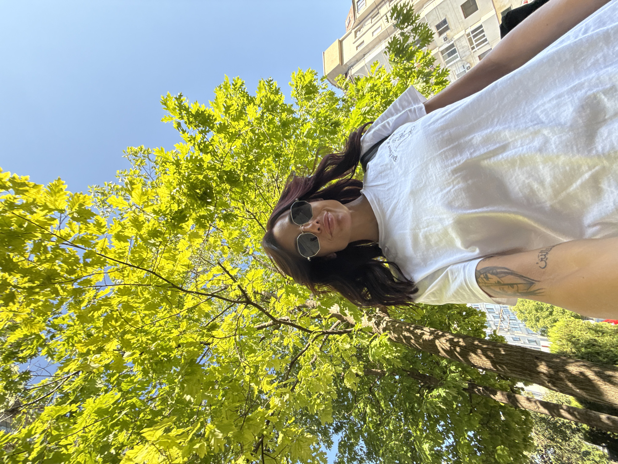 A low-angle, wide shot shows a woman taking a selfie outdoors in Pleven, Bulgaria. She is wearing sunglasses and a white t-shirt. The woman is standing in front of a large, leafy tree with bright green foliage. The sky is clear and blue. In the background, a multi-story building with visible windows and air conditioning units is partially visible. The woman has a tattoo on her arm, partially visible. The image is shot during the daytime, with sunlight illuminating the scene.
