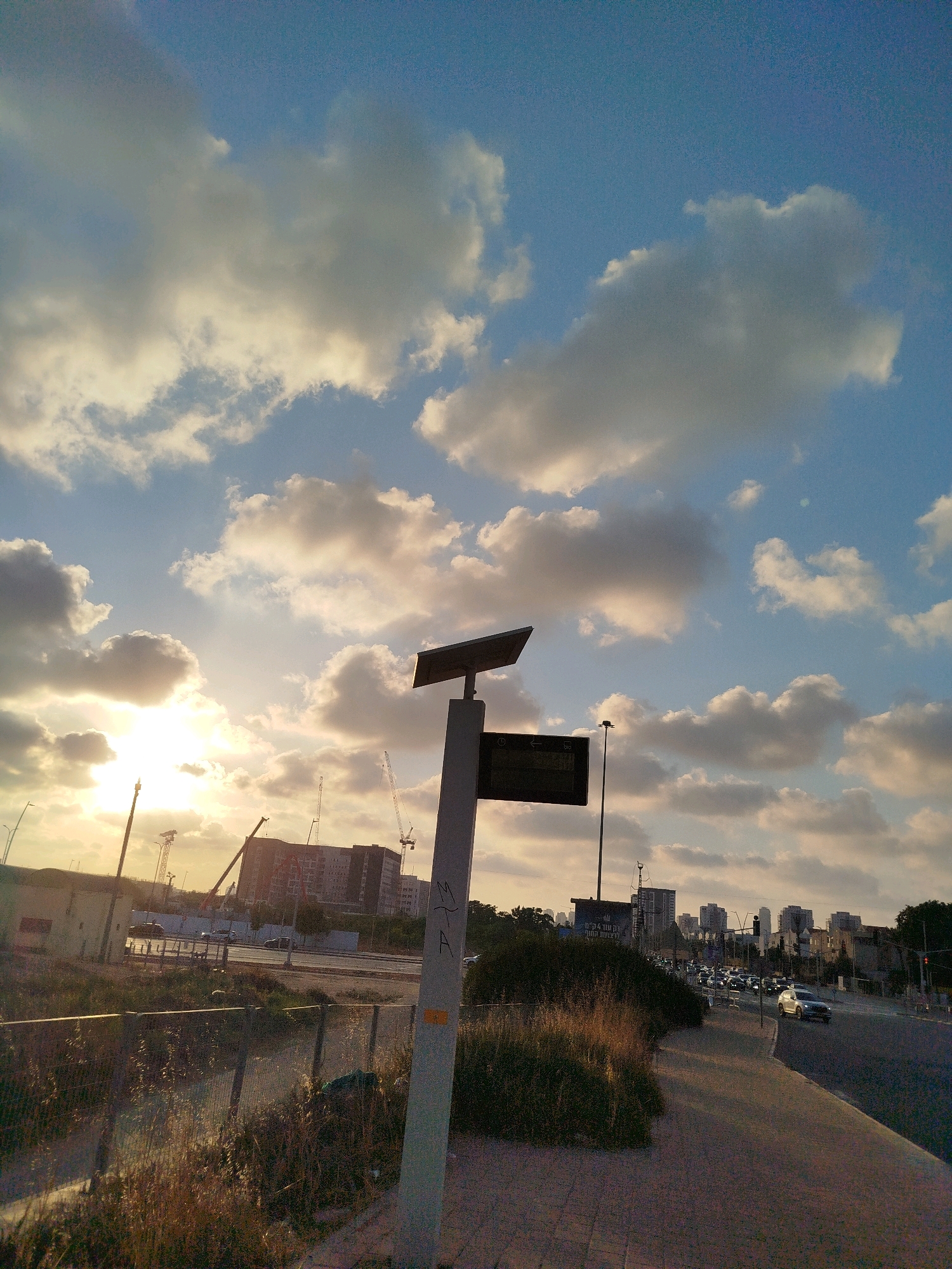 This is a vertical, low-angle, eye-level shot of a street scene in Ashdod, Israel, at sunset. The sky is a gradient of blue and orange, filled with soft, fluffy white clouds. The sun, partially obscured by clouds, casts a warm glow on the scene. In the foreground, a sidewalk with a checkered pattern is visible on the right, leading to a road with cars driving in both directions. A green bush with dry grass is on the left side of the sidewalk. A tall, thin pole with a solar panel on top stands in the middle of the frame, with a dark rectangular sign attached to it. The sign contains Hebrew text. Further in the background, there are buildings, construction cranes, and streetlights, indicating an urban environment. The overall mood is peaceful and serene, with a touch of urban hustle.