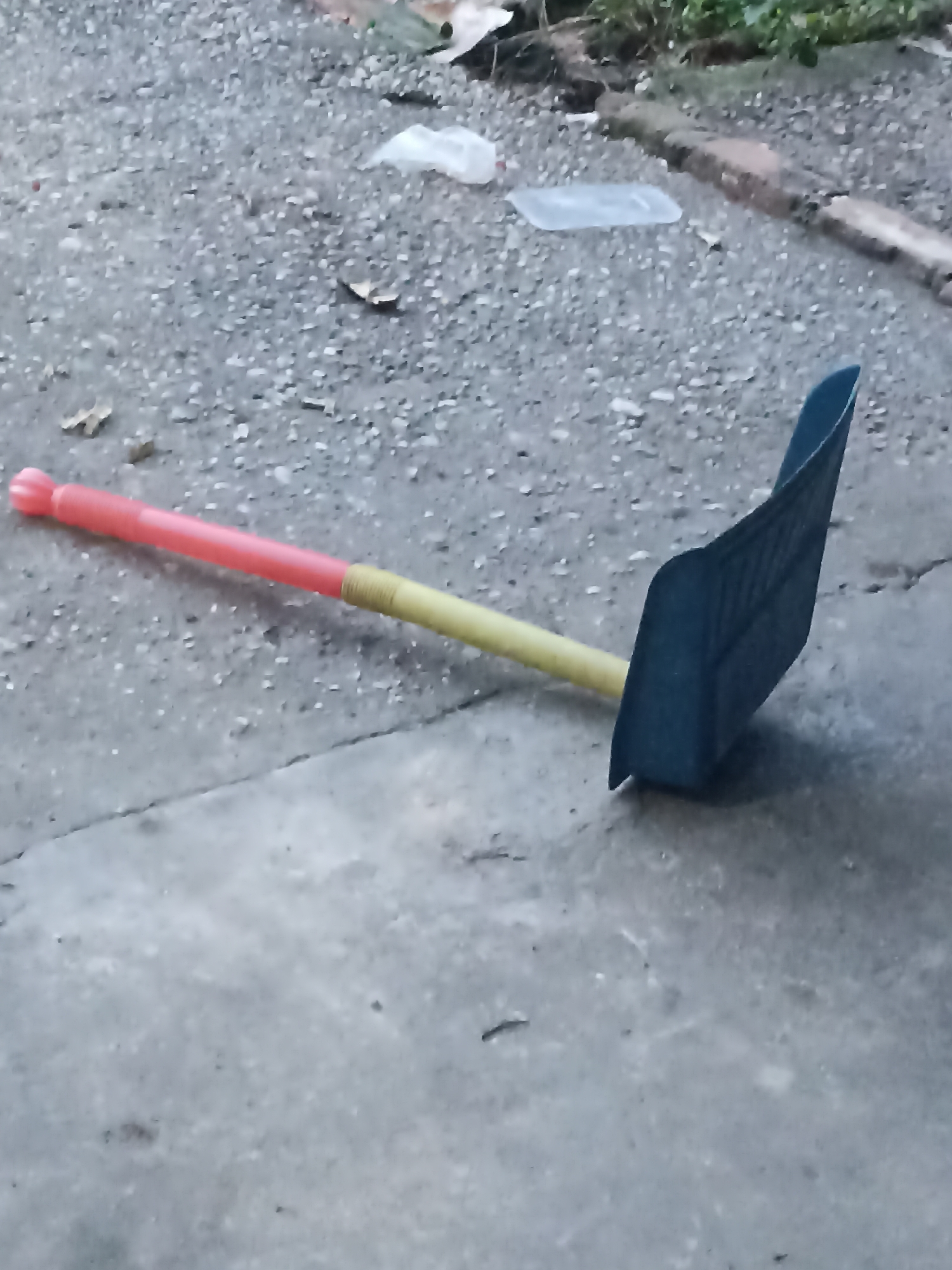 A blue dustpan with a red and yellow handle lies on an asphalt surface. The dustpan is positioned to the right of the frame, with its opening facing upwards and slightly to the left. The handle extends to the left, with a red grip at the far end and a yellow section leading to the blue dustpan. The asphalt surface appears to be part of a pathway or street. In the background, there is debris scattered on the ground, including leaves, plastic bags, and a discarded plastic container. Some vegetation is visible in the upper right corner. The overall scene depicts an outdoor environment that is somewhat neglected.