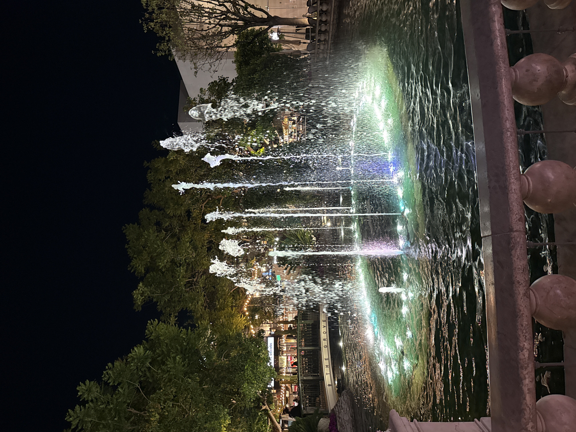 The image shows a nighttime view of the fountain at The Grove, an outdoor shopping and entertainment complex in Los Angeles, United States of America. The fountain is actively spraying water, and the water is illuminated with colorful lights. In the background, the architecture of The Grove is visible, including storefronts and pedestrian areas, lit by ambient lights. The foreground includes a portion of the fountain's edge and decorative railings. The sky is dark. Trees are present in the mid-ground, flanking the view of the fountain and buildings. People are visible in the background near the storefronts.