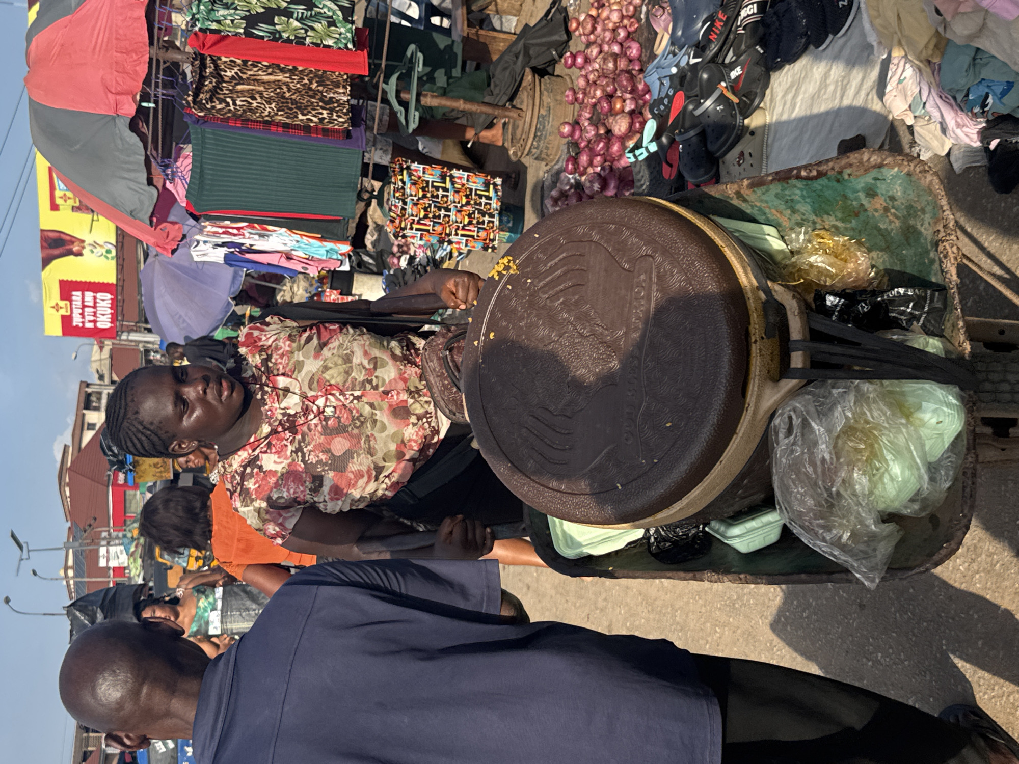 The image depicts a bustling market scene in Onitsha, Nigeria, bathed in bright sunlight. In the foreground, a woman with braided hair, wearing a floral print blouse, stands behind a wheelbarrow filled with items, including a large brown container with a textured lid, plastic bags containing green items, and other miscellaneous objects. Her left hand grips the wheelbarrow's handle. To the left of the woman, a man in a dark blue shirt and black pants is partially visible, facing away from the camera. The wheelbarrow itself is worn and appears to be made of metal with green paint.
Behind the woman, a variety of market stalls are set up. Clothing items, including skirts with various patterns like leopard print and green ribbed fabric, are displayed on hangers under a red and gray umbrella. Further back, more clothing hangs, and various goods are piled on the ground. A large billboard with red and yellow branding is visible in the background, advertising "JUAPUTARA N'UTO ANU OKUKO." Numerous people can be seen moving through the market, some interacting with vendors, while others are simply passing by. A collection of red onions is displayed on the ground near a pile of footwear, including sandals and clogs. The ground surface is paved, and the sky above is clear blue. The overall impression is one of vibrant commercial activity in an open-air market.