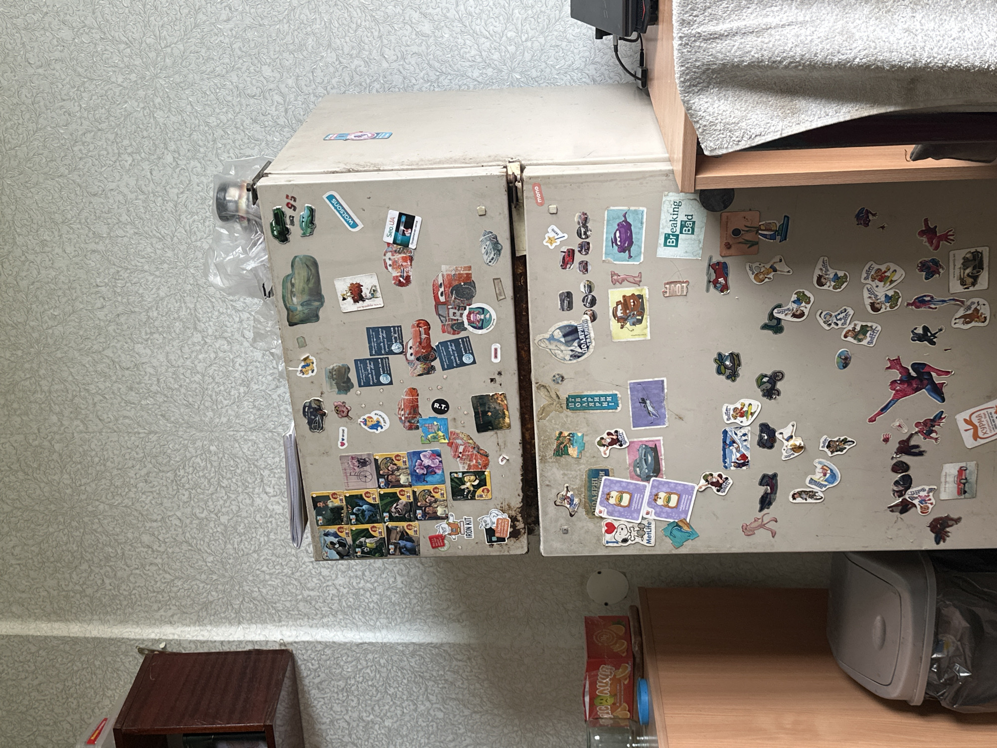 The image shows a close-up of a two-door refrigerator covered in various stickers, located in a room with patterned wallpaper. The refrigerator is a light beige color and appears to be older, with some visible rust spots. The stickers include cartoon characters, movie posters, and miscellaneous graphics. On the top door, there are stickers featuring characters from the movie "Cars," a "Watsons" logo, and other animated figures. The bottom door displays stickers of "Snoopy," "MetLife" branding, characters from "Breaking Bad," and more animated images. To the left of the refrigerator, there is a wooden cabinet with a red box of what appears to be tea or snacks on top, and a gray trash can with a black trash bag. To the right of the refrigerator, a portion of a wooden countertop is visible with a black electronic device. The overall environment appears to be a residential kitchen or utility area. No people are visible in the image. The specific location in Kyiv, Ukraine cannot be determined from the visual information provided.
