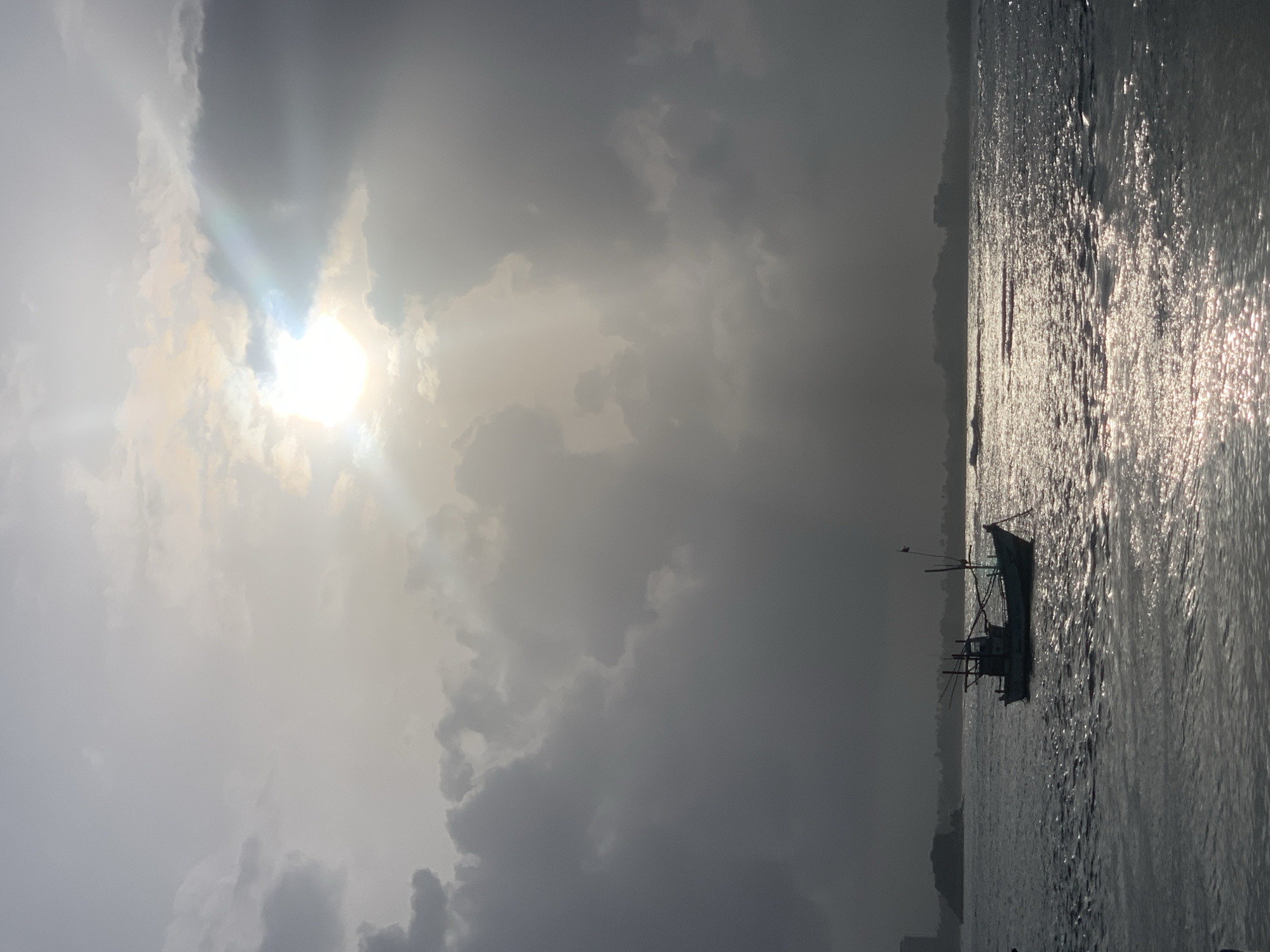 A lone fishing boat is silhouetted against the shimmering water of the ocean. The sun is partially obscured by dramatic clouds, casting rays of light that create a dramatic contrast between light and shadow. In the distance, the coastline is visible with a few buildings. The scene captures the raw beauty of the ocean, the power of nature, and the solitude of the sea.