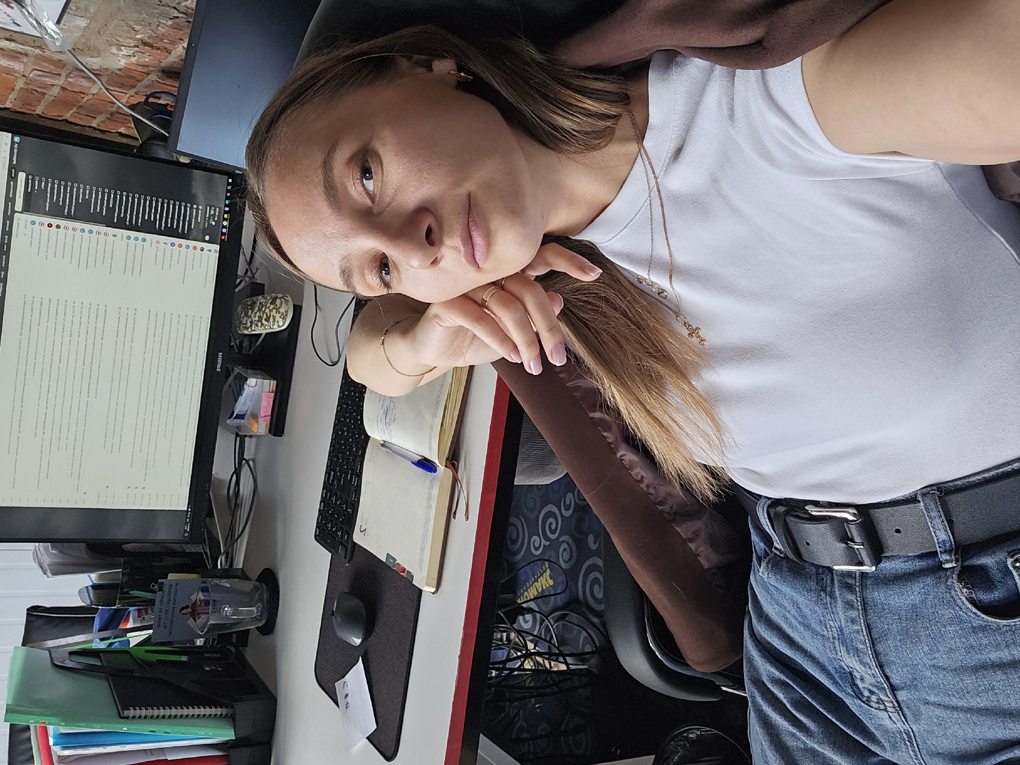 The image shows a woman with light brown hair wearing a white t-shirt, a gold necklace, and blue jeans with a black belt. She is sitting at a desk with a computer monitor, keyboard, mouse, notebooks, pens, and other office supplies. The monitor displays a text-heavy interface. The location is possibly an office setting in the city of Chaikovsky, Russia. The woman is resting her head on her hand, and the background includes a brick wall. The environment suggests an indoor workspace.