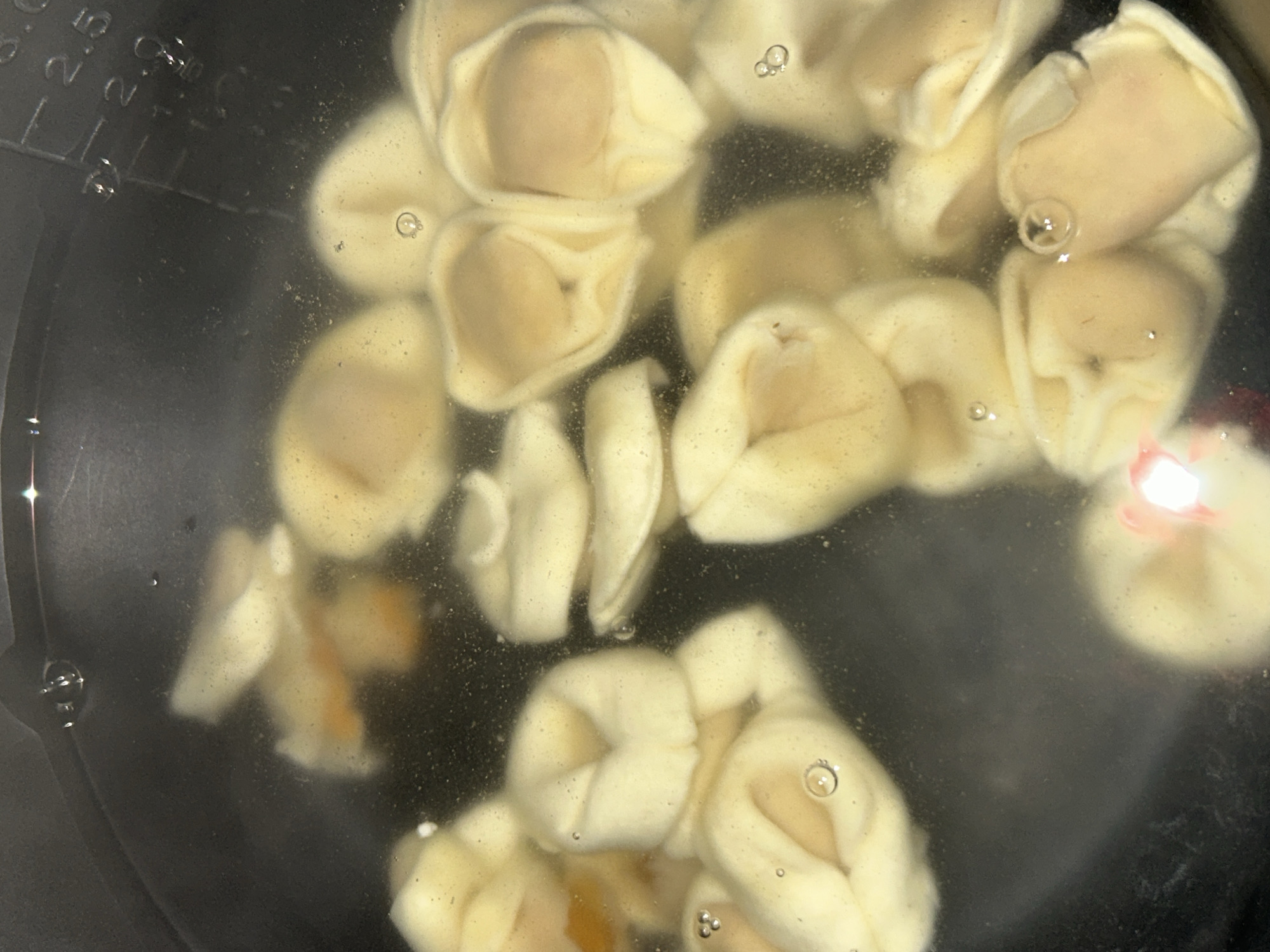 A close-up, overhead shot shows several pieces of tortellini pasta submerged in clear liquid, likely water, within a dark pot. The tortellini are pale yellow and appear to be cooking, as evidenced by small bubbles rising to the surface. Some tortellini are partially obscured by others. The pot has subtle measuring marks visible on the upper right edge, indicating measurements up to 3.0. The lighting is somewhat dim, with a bright reflection on the left side of the image, possibly from a light source. There are no people or animals present in the image. The scene depicts food preparation. The image does not contain information that would allow for the determination of its location within the city of Sofia, Bulgaria.