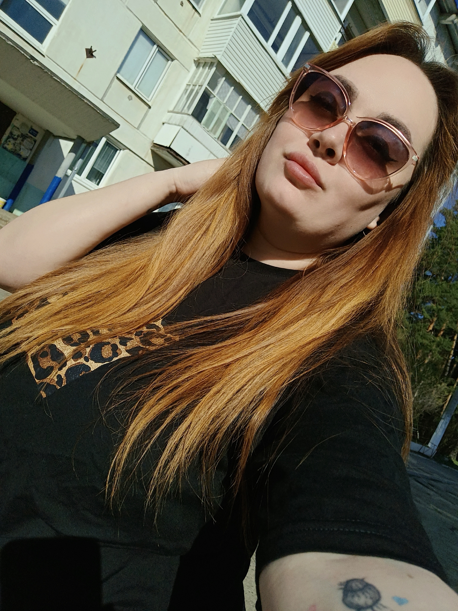 A woman is taking a selfie outdoors in Chaikovsky, Russia. She has long, light brown hair and is wearing sunglasses. She is wearing a black t-shirt with a leopard print design. A multi-story building is behind her to the left and trees are to the right. She has a tattoo on her left arm.