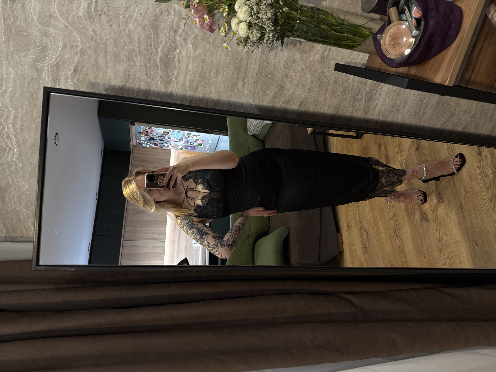 A reflection in a rectangular mirror reveals a woman with blonde hair, wearing a black dress with lace detailing, holding a phone in front of her face. Her left arm is extensively tattooed. She is standing in what appears to be an apartment or house, with a visible interior including a green chair and dark furniture. The setting includes a side table with a flower arrangement and makeup items. A brown curtain is partially visible at the bottom of the mirror, suggesting the bottom of the mirror's support structure. Based on the available visual context and without specific landmarks, it is impossible to confirm the location is in Krasnoyarsk, Russia.