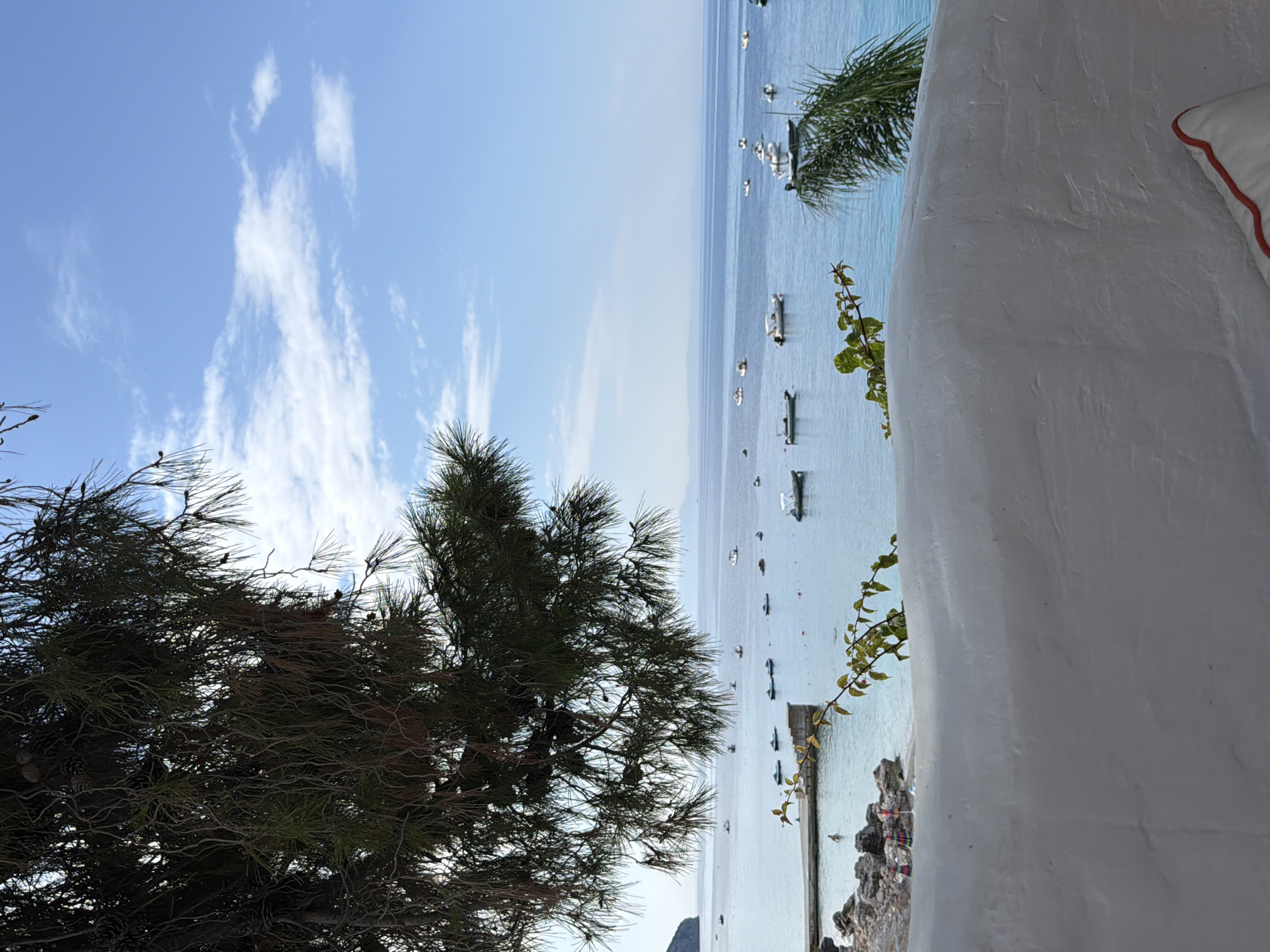 A view of the sea in Himarë, Albania, is framed by a pine tree in the foreground and a white stucco wall with a red-trimmed pillow. The calm, turquoise water is dotted with numerous small boats. A concrete pier extends into the water on the left side of the image, where a single person is swimming. A small, rocky beach with a few umbrellas is visible near the pier. The sky is a clear blue with scattered white clouds, and distant mountains are faintly visible on the horizon. The overall impression is one of a peaceful, sunny day at the seaside.