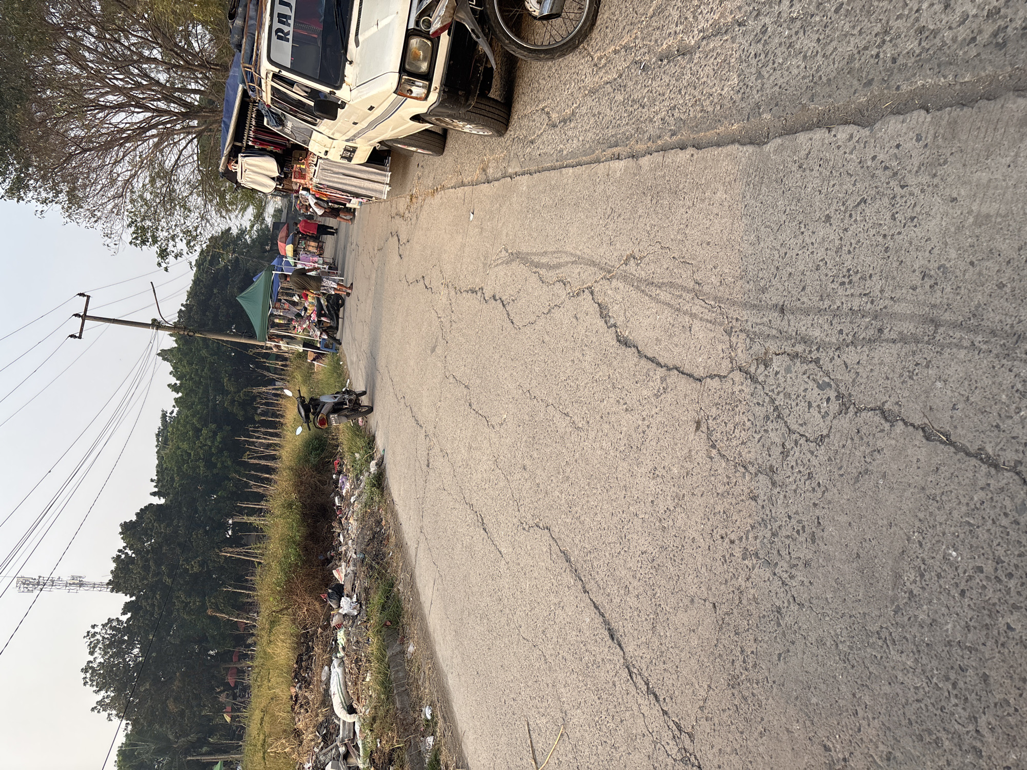 The image depicts a street scene in Telagamurni, Indonesia, characterized by a roadside market and some evidence of urban disarray. In the foreground, a cracked asphalt road dominates the lower portion of the frame, showing signs of wear and tear. To the right of the road, a white pickup truck is parked, displaying the word "RAJA" on its side. Next to the truck, market stalls are set up, with various goods, including clothing, displayed on racks. Several individuals are visible browsing or tending to the stalls. Further down the road, more market tents and people are present, suggesting an active commercial area. To the left of the road, a strip of land is covered in trash and debris, with a motorcycle parked nearby. In the background, lush green trees form a backdrop to the scene, with utility poles and power lines extending across the sky. The overall atmosphere suggests a bustling but somewhat unkempt urban environment.