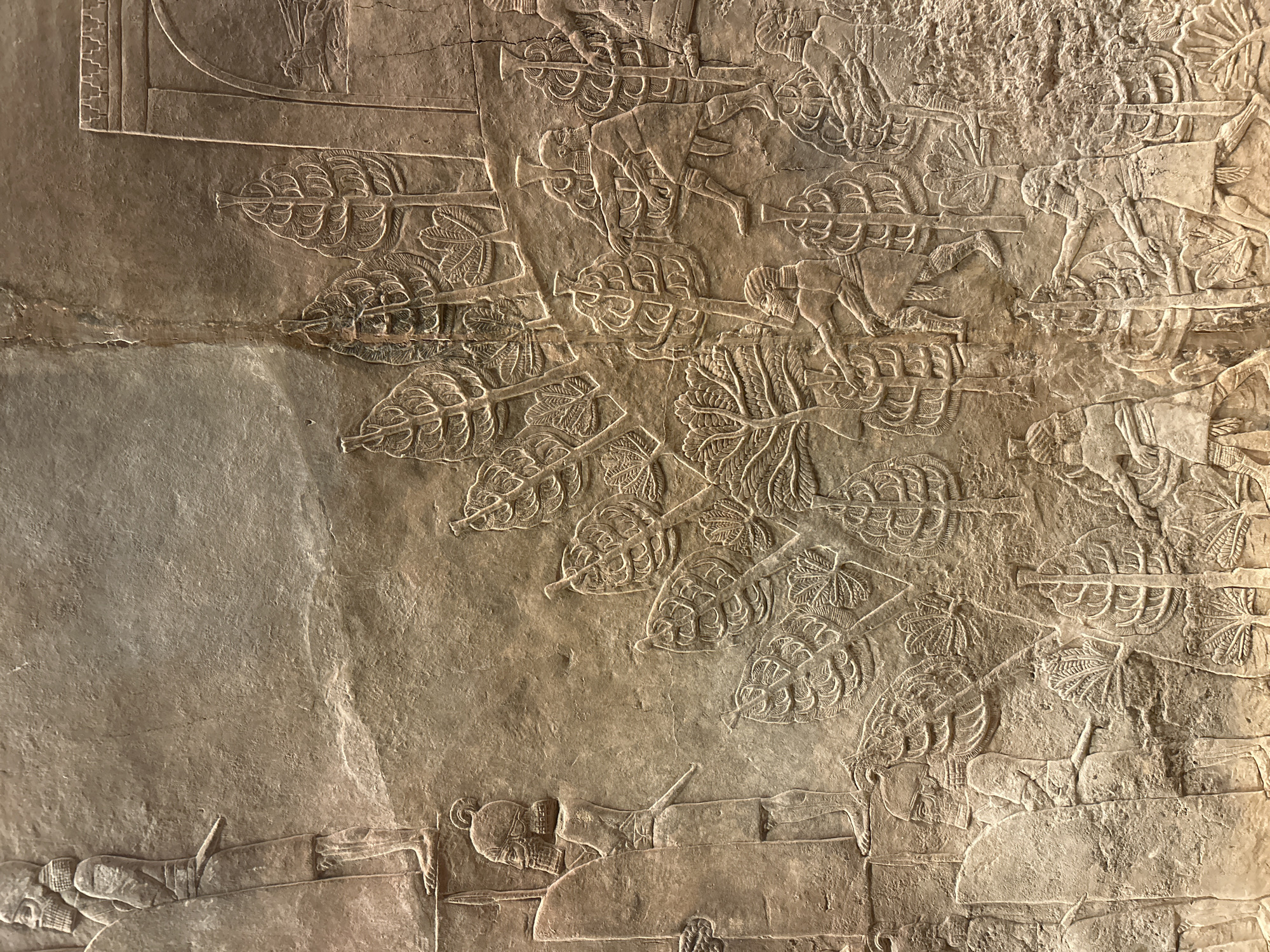 This is a close-up photograph of a section of a bas-relief carving on a stone wall, likely located in the British Museum, London, United Kingdom. The relief depicts a scene with several figures, possibly soldiers or attendants, interspersed with stylized trees. The figures are carved in profile, with detailed beards and hair, wearing segmented skirts or robes and some form of headdress. Some figures are shown carrying spears and shields. The trees are depicted with rounded, layered foliage and segmented trunks. The carving is ancient, characterized by its textured stone surface and the worn appearance of the relief. The overall impression is one of a historical or mythological narrative rendered in stone.