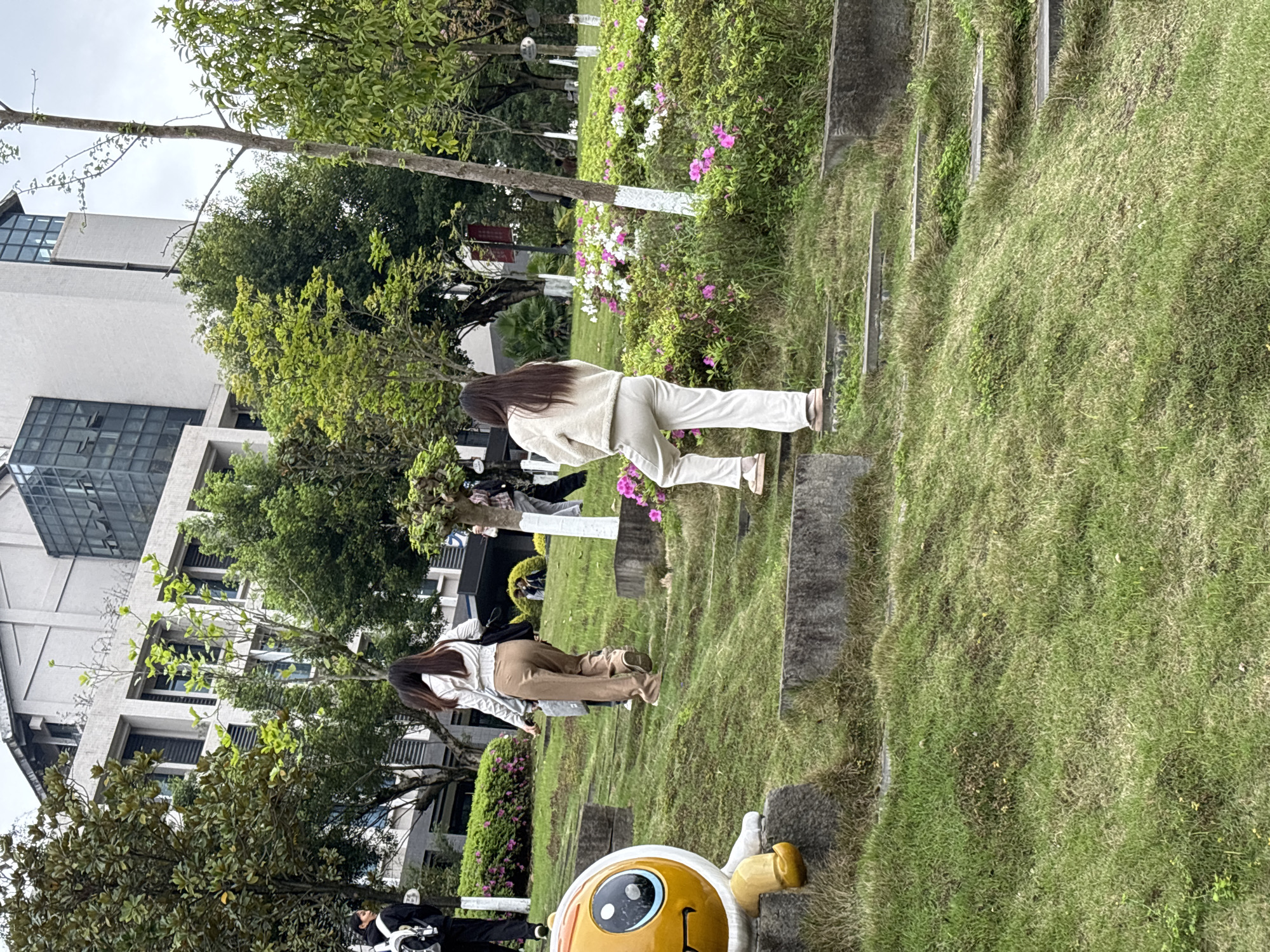 The image depicts an outdoor scene in what appears to be a park or garden setting in Jiangjin, China. There are two women walking in the park or garden among lush green grass, trees, and flowering shrubs. There is an animated statue in the foreground, seemingly part of the park’s decor. A large, multi-story building stands in the background.