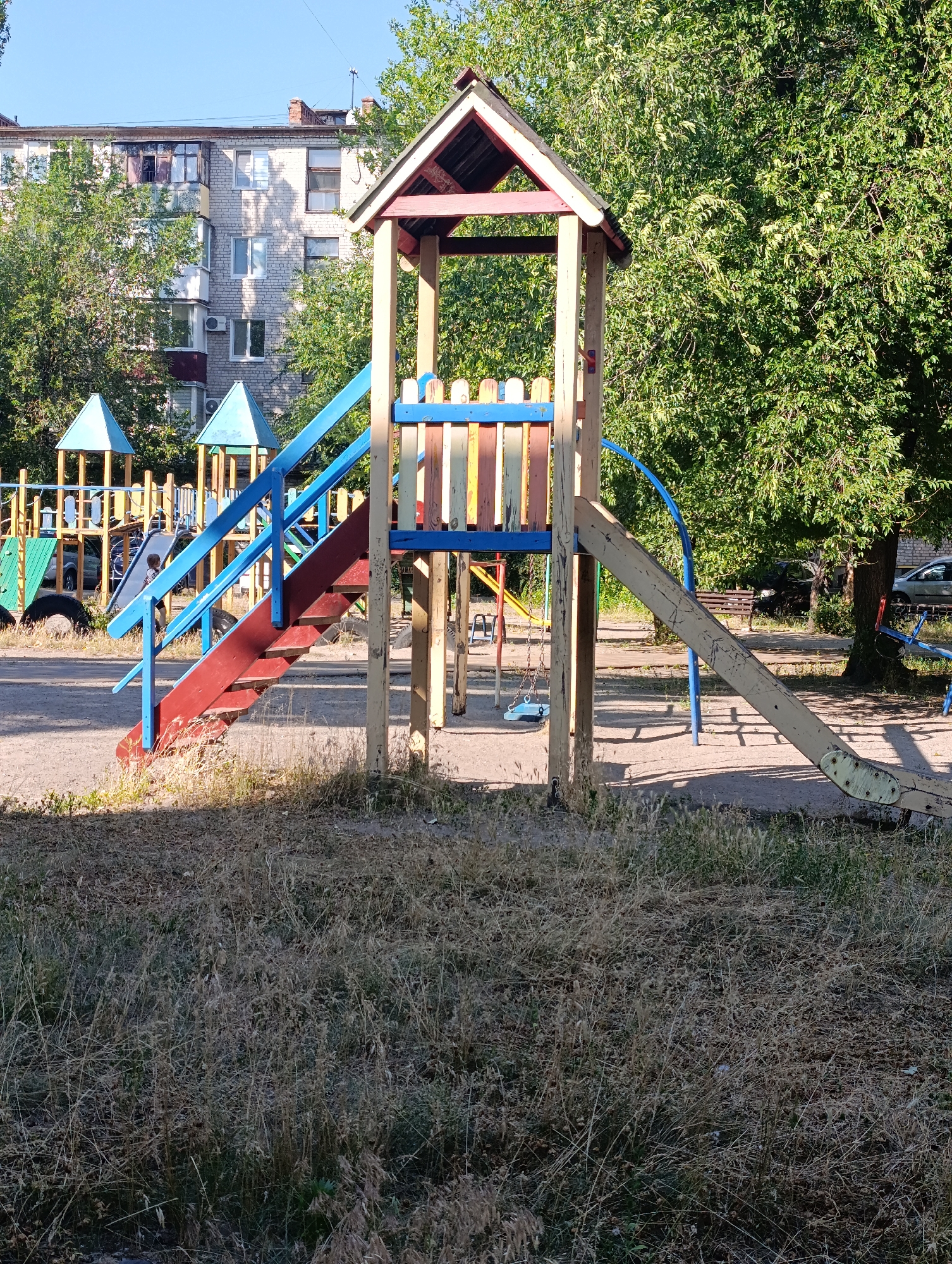 The image depicts a children's playground in an urban setting, bathed in the bright light of a sunny day. The playground features a wooden play structure with a slide, stairs, and colorful railings. Behind the play structure, a multi-story apartment building is visible, suggesting a residential area. Lush green trees surround the playground, providing shade and a natural element to the scene. The ground is covered with sand, typical of playgrounds, and dry, overgrown grass is present in the foreground. Other playground equipment, including swings and a seesaw, can be seen in the background. The overall impression is one of a quiet, possibly slightly neglected, public space on a clear day. There is no visible text in the image.