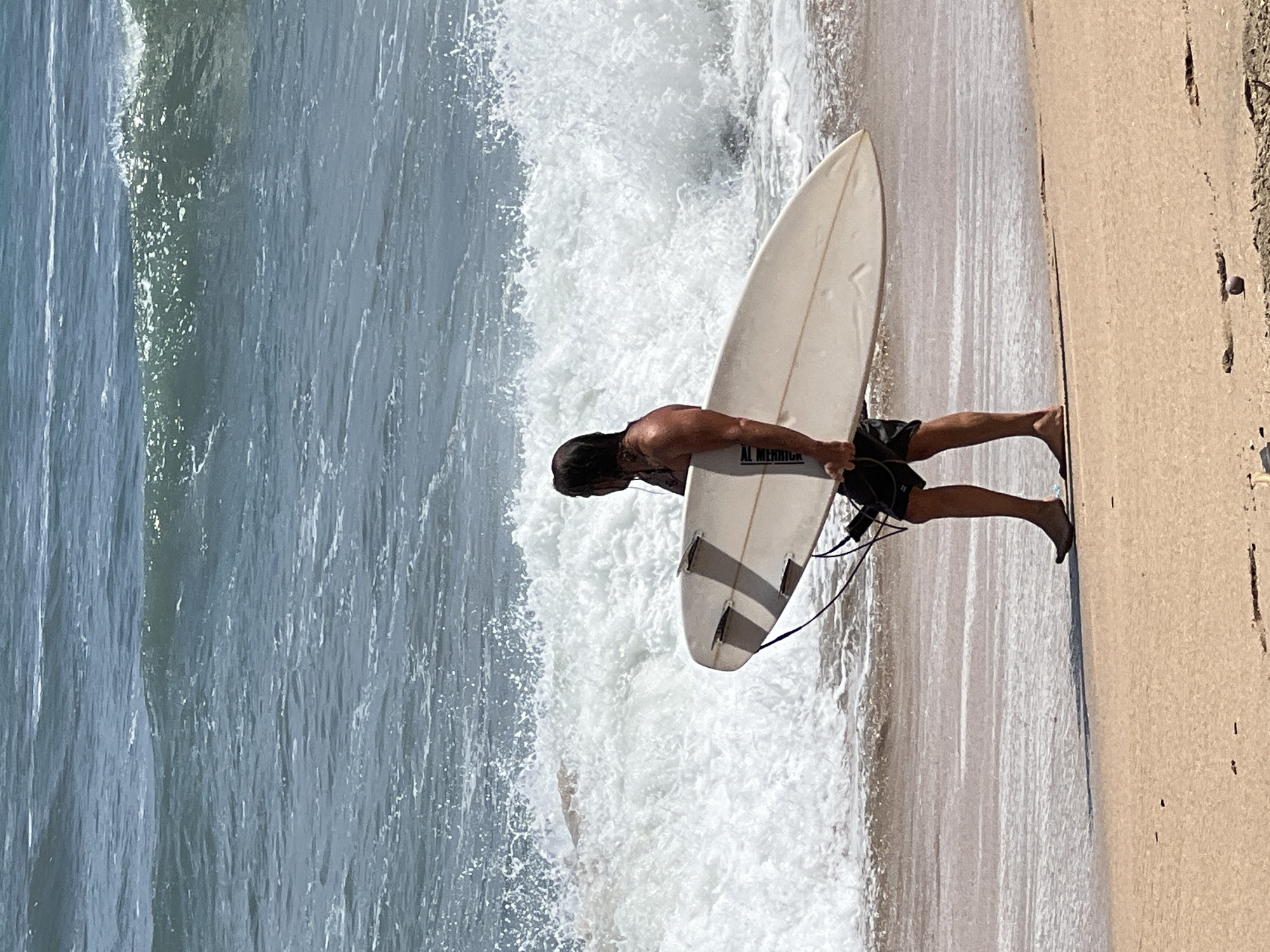 A surfer walks along the beach in Kuta, Indonesia, carrying a surfboard. The surfer, seen from the side, has long dark hair and is wearing dark swim trunks. The surfboard is white with three fins on the bottom and a leash attached. The ocean behind the surfer is rough with white-capped waves breaking. The beach is sandy and appears to be slightly wet near the water's edge. The sun is shining, casting shadows on the sand.