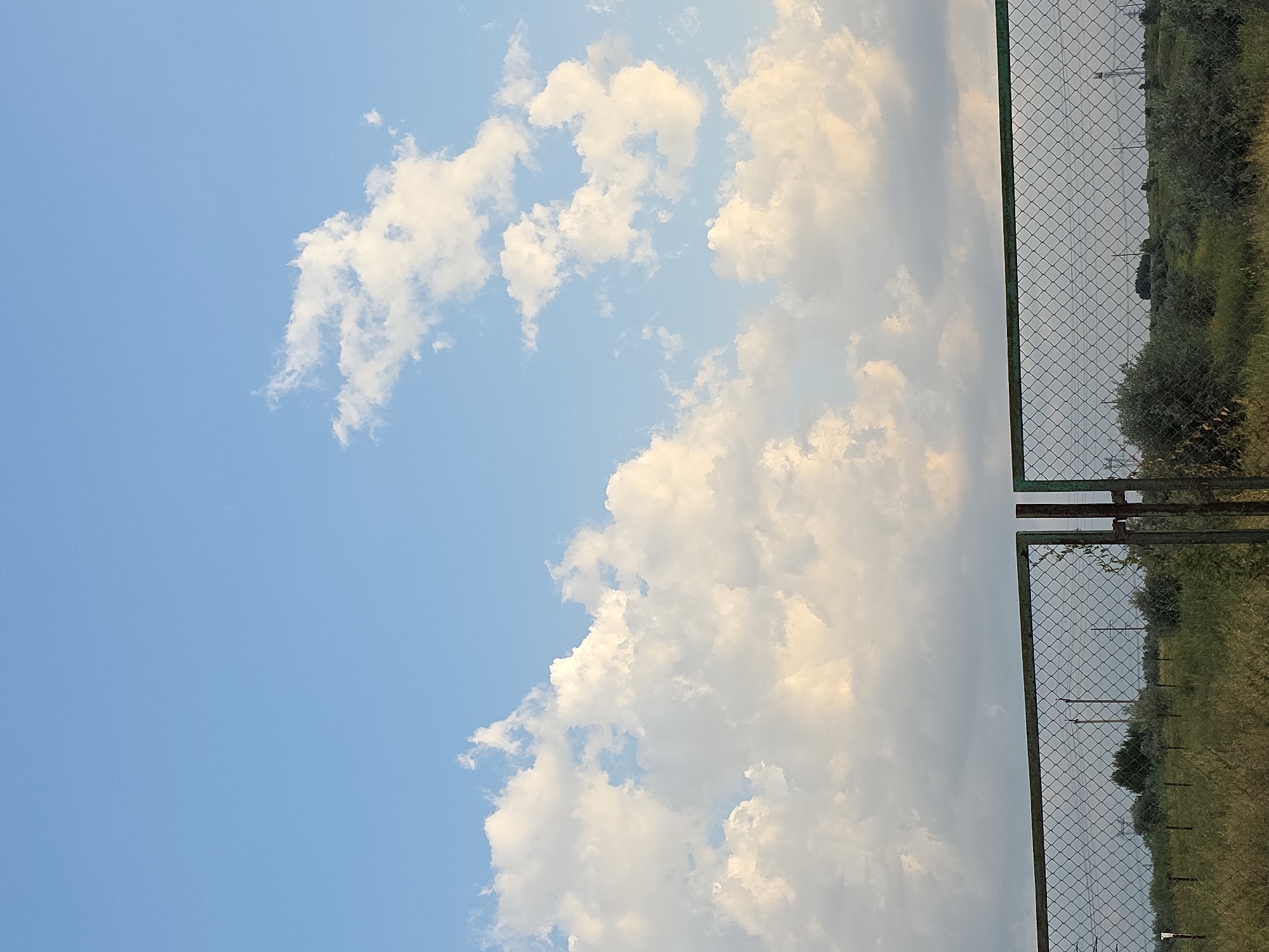 This image depicts a chain-link fence in the foreground, with a green metal frame and rust on its edges. The fence is approximately two panels wide and appears to be open in the center, suggesting a gate or an opening. Beyond the fence, a flat, grassy landscape stretches to the horizon, dotted with sparse trees and bushes. Several utility poles and power lines are visible in the distance, indicating some infrastructure in the area. The sky above is a vibrant blue, filled with scattered, fluffy white clouds, some of which are tinged with yellow from the sunlight. The overall scene is open and rural, likely situated in Podgorodka, Kazakhstan, given the context. There are no people or animals present in the image. The lighting suggests it is daytime, possibly late afternoon or early evening due to the warm tones on some of the clouds.