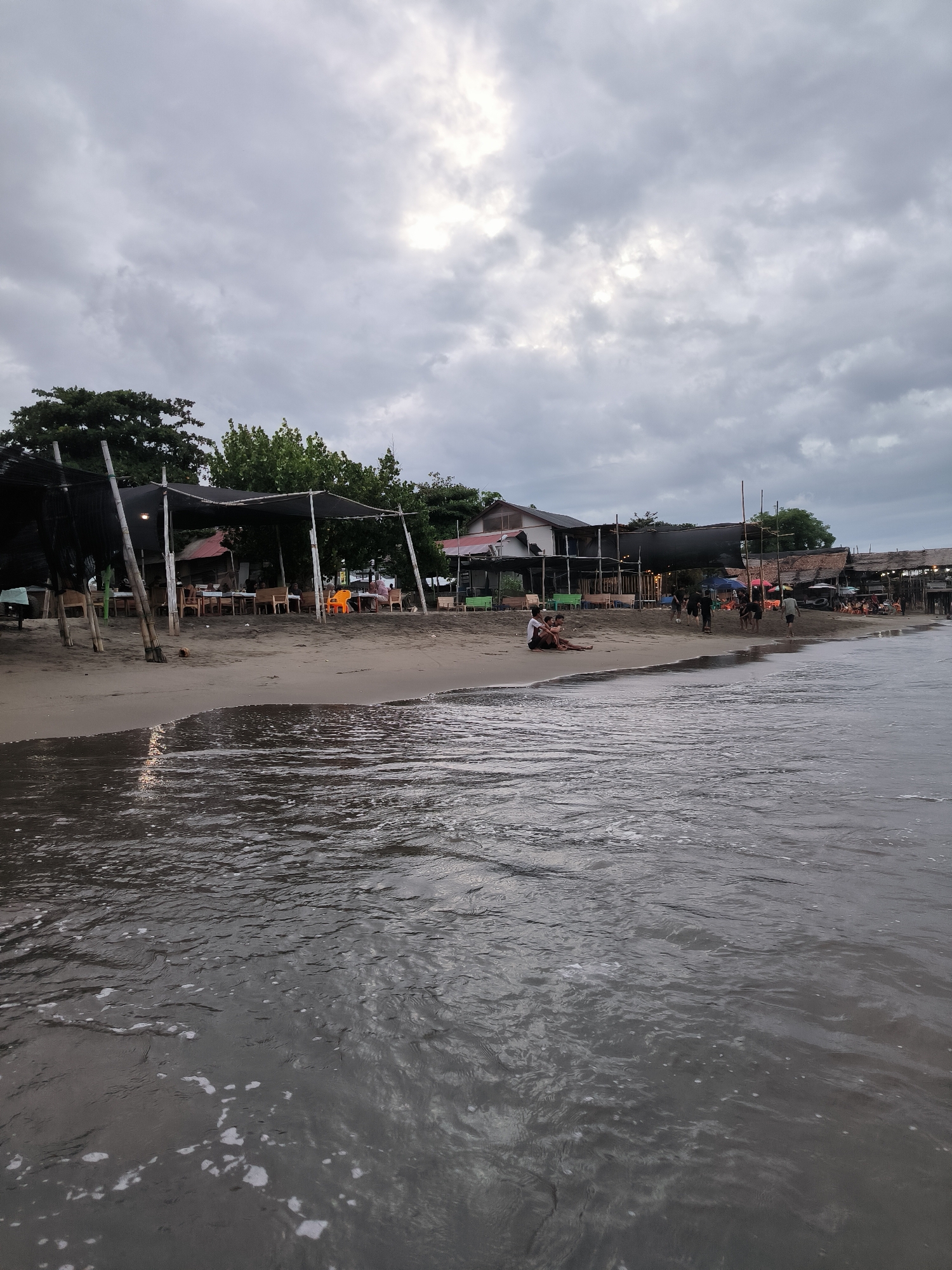 This image captures a wide-angle view of a coastal scene in Lhokseumawe, Indonesia, under an overcast sky. The foreground and right half of the image are dominated by dark, rippled ocean water with intermittent patches of white foam. The water's surface reflects faint, lighter tones from the sky.
A dark sandy beach occupies the left side of the foreground and extends into the mid-ground. Along this beach, a series of informal, open-air structures, likely dining establishments, are erected. These structures feature dark, possibly tarp or thatched, roofs supported by wooden poles. Underneath these coverings, multiple tables and chairs (including one distinct orange plastic chair) are arranged, with several individuals seated, appearing to be dining or socializing. String lights are visible within some of these structures.
Behind the structures, dense green foliage from various trees is present, with a few larger buildings discernible further in the background on the left.
On the beach itself, in the mid-ground left, two individuals are seated directly on the sand near the water's edge, facing the ocean. Further along the beach to the right, a group of approximately ten individuals is walking on the sand, while another person is partially submerged in the shallow water near the shore. A single coconut rests on the sand near one of the covered seating areas. The overall light condition suggests late afternoon or an overcast day.