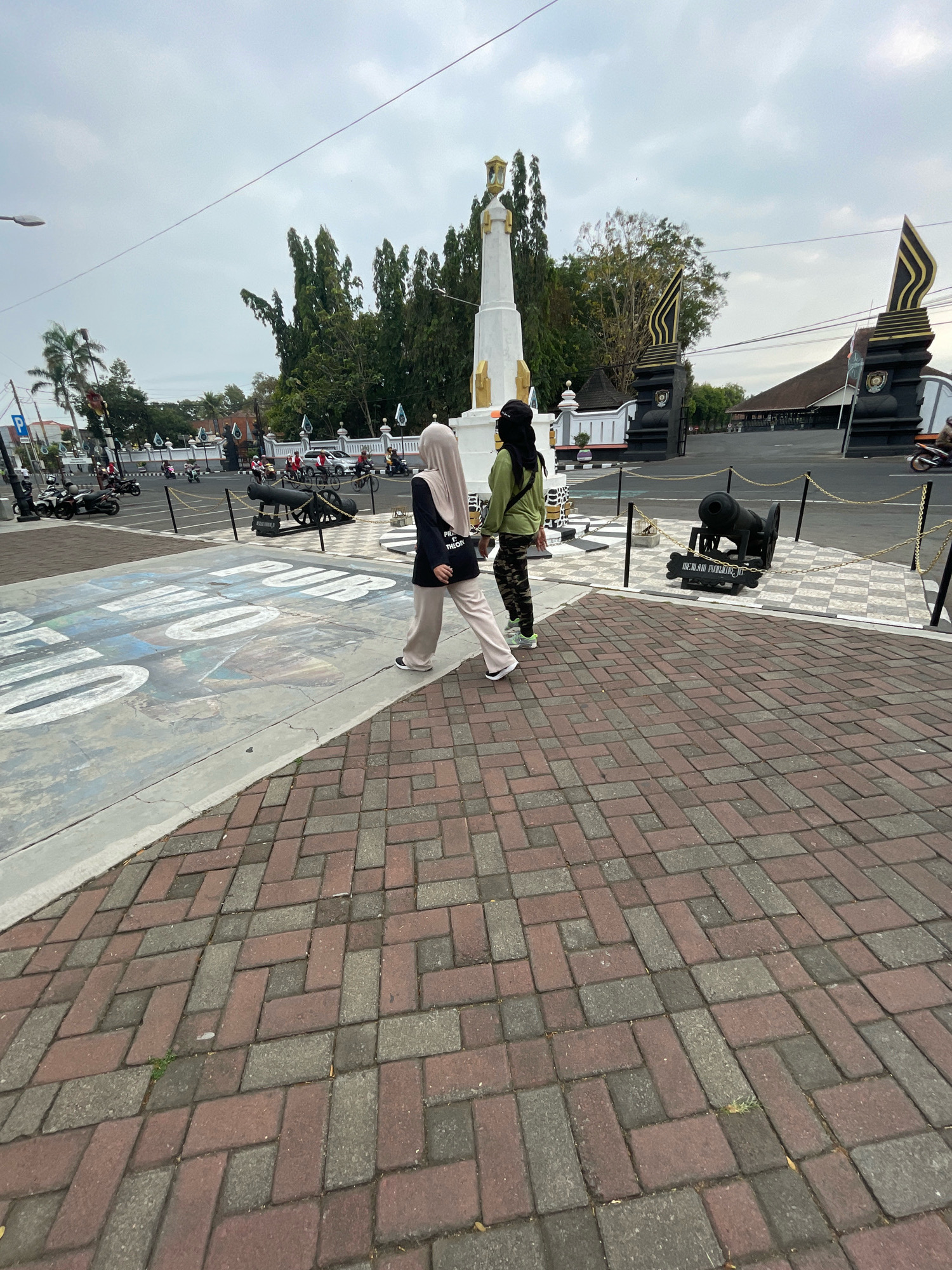 The image captures an outdoor scene in the city of Purworejo, Indonesia, likely near a historical monument. In the foreground, two individuals, both appearing to be women, are walking away from the camera on a paved area. One is wearing a light brown hijab and light-colored pants, carrying a black bag with text. The other is wearing a dark headscarf and camouflage-patterned pants.
Behind them, a large white monument with gold accents stands prominently. The monument is flanked by decorative black pillars with gold finials. In the middle ground, several old cannons are displayed on raised platforms, surrounded by yellow chains and checkered pavement. Beyond the monument and cannons, a street is visible with traffic, including motorcycles and cars. Palm trees and other lush green trees form a backdrop, with a cloudy sky overhead. The ground in the foreground is paved with reddish-brown and gray bricks laid in a herringbone pattern. A large painted mural is visible on a concrete surface to the left of the foreground paving. The overall impression is that of a public space commemorating historical events.