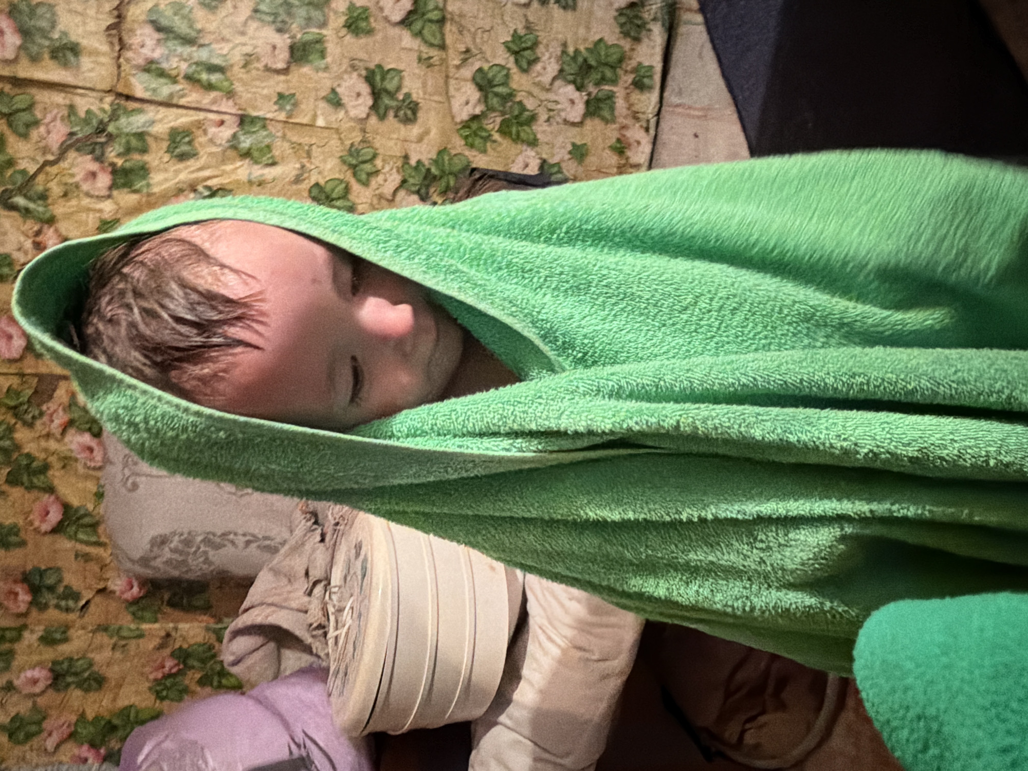 A toddler, wrapped in a bright green towel, stands in what appears to be a room with floral wallpaper. The child's head is slightly bowed, and their wet hair is visible under the towel's hood. The background shows pillows and a round, tiered container, suggesting a home environment. There is no discernible location information within the image to associate it with Krasny Mayak, Russia.