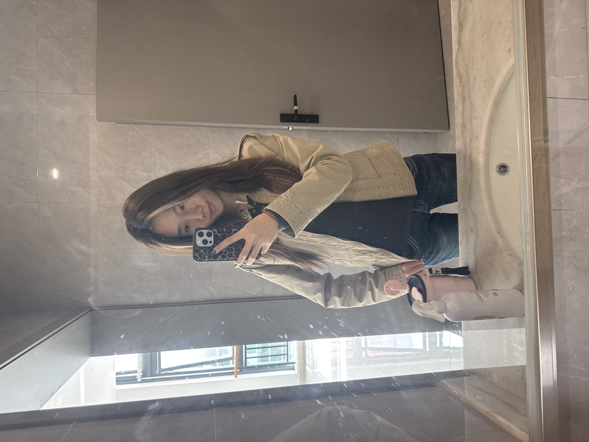 A young woman is taking a selfie in a bathroom mirror. She is wearing a beige jacket, a black top, and blue jeans. She is holding a phone with a leopard print case. On the counter next to the sink, there is a pink water bottle and a white bottle with a spray nozzle. The bathroom has light grey tile walls. The reflection in the mirror shows a window with a view of a building in Nan'an District, China.