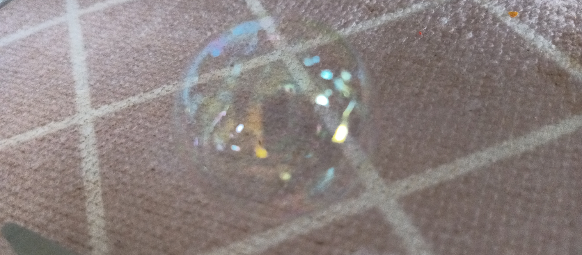 A soap bubble rests on a tan surface patterned with white lines forming a grid. The bubble exhibits a spectrum of colors reflected in its thin film, including iridescent blues, yellows, and greens. The surface beneath is textured, with a fine granular appearance.