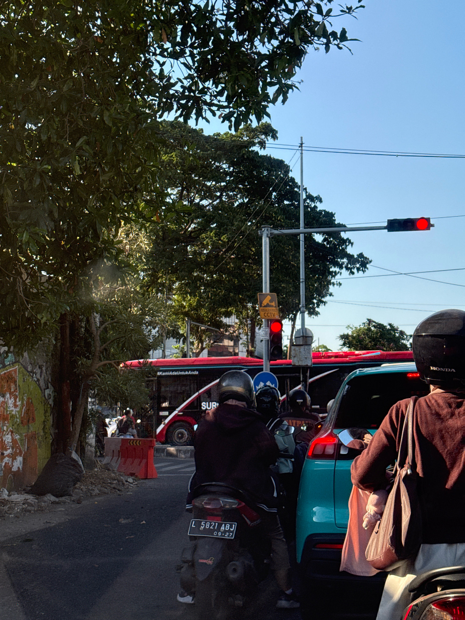 The image depicts a traffic scene in Surabaya, Indonesia. In the foreground, the back of a scooter is visible with a license plate reading "L 5821 ABJ" and the date "09-27". Behind the scooter, a turquoise car is stopped, and a person wearing a dark brown long-sleeved shirt and a bag slung over their shoulder is partially visible in the driver's seat. A large red bus with the word "SURABAYA" visible on its side is stopped in traffic ahead. To the left of the bus, a traffic light shows a red signal. A CCTV sign is also visible on a pole. The scene is illuminated by sunlight, casting shadows on the road. Trees line the side of the road, and a clear blue sky is visible above.