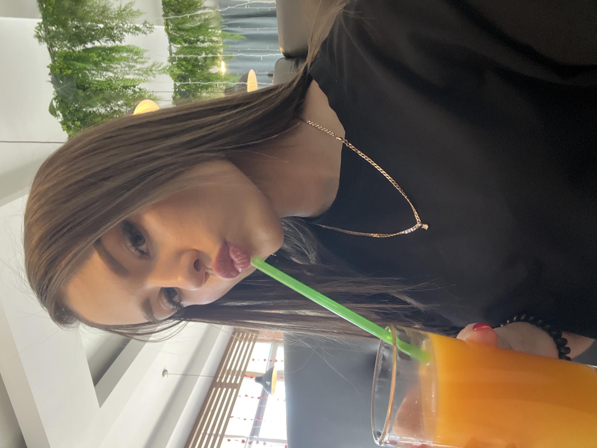 A young adult woman with brown hair is drinking orange juice through a green straw in a restaurant or cafe. She is wearing a black shirt and a delicate gold necklace. Her nails are painted red. The background shows a restaurant interior with greenery and a window. Based on the window decorations featuring a heart motif, the location is in the city of Chaikovsky, Russia, due to the city's known Valentine's Day decorations in public spaces.
