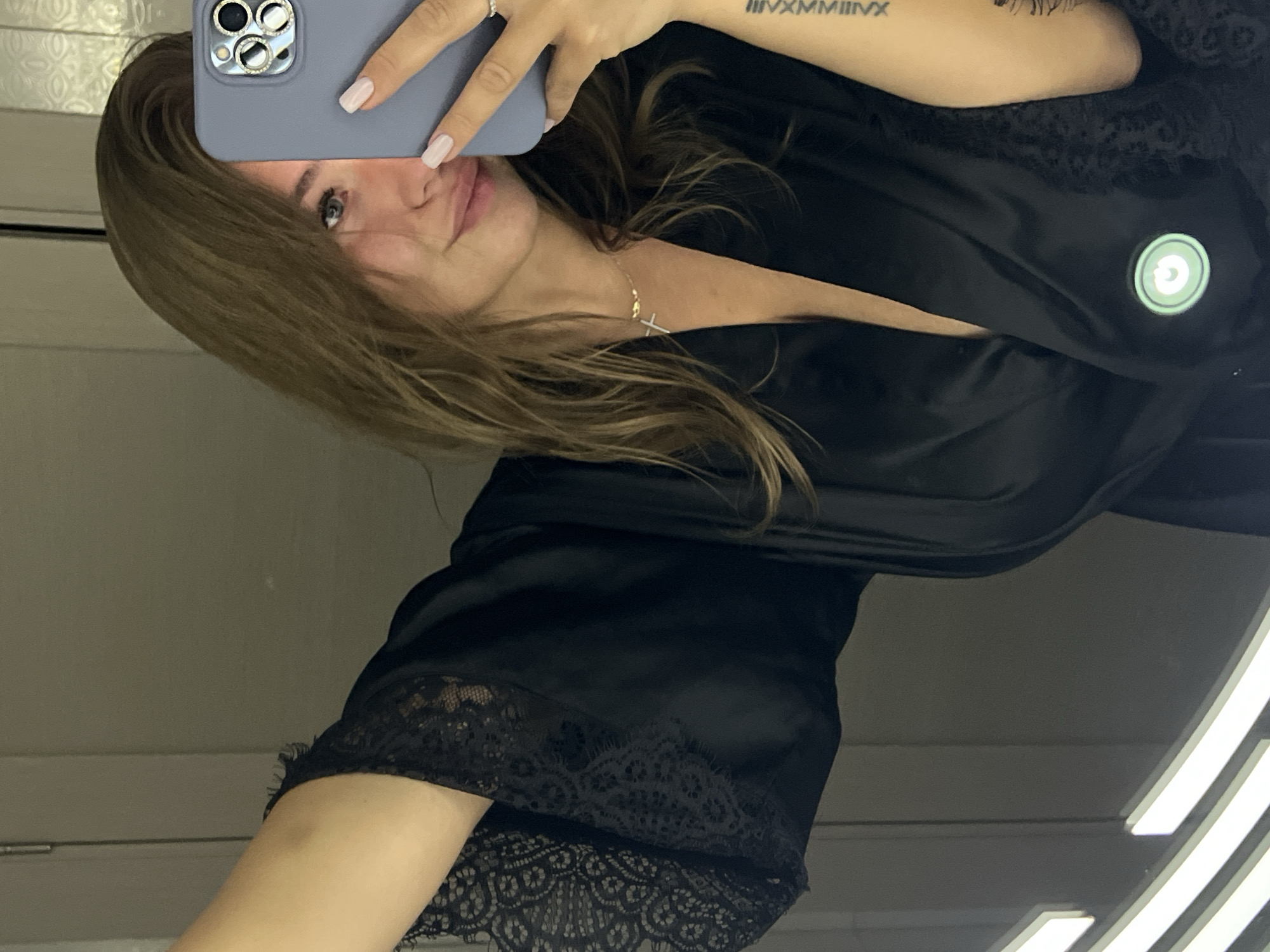 A woman takes a selfie in what appears to be a changing room or dressing room. She is wearing a black robe with lace trim. She is holding a gray smartphone in her left hand, partially obscuring her face. Her hair is brown and partially covers her face. A gold necklace with a cross pendant is visible. A tattoo is visible on her left arm. The background shows a light gray wall and fluorescent lighting. There is no clear indication of the location being in Budva, Montenegro.