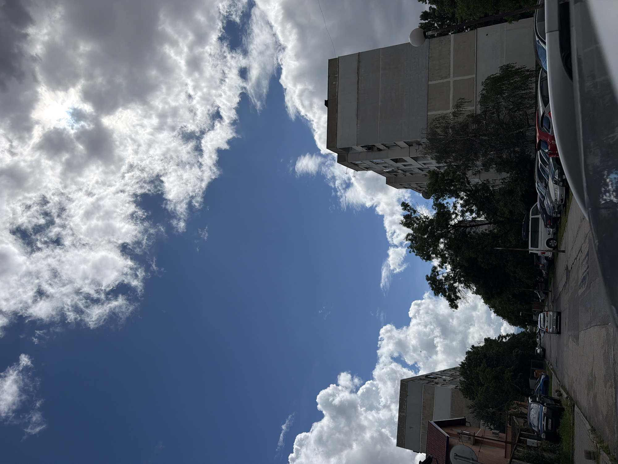 The image captures a daytime street scene in Sofia, Bulgaria, on a partly cloudy day, viewed from a low angle looking upwards. The upper left of the frame is dominated by a bright blue sky with a mix of large, fluffy white cumulus clouds and thinner, wispy clouds. The sun, intensely bright, is partially obscured by clouds in the upper left, creating a radiant highlight.
On the right side of the image, the upper portion features a tall, light-grey multi-story apartment building with a flat roof, visible windows, and balconies. Below it, a narrower street runs roughly horizontally, lined with numerous parked cars of various makes and colors, including white, grey, red, and black vehicles. Further down the street, another building with a reddish-brown tiled roof and a white satellite dish on its facade is partially visible.
Dense green trees are scattered throughout the scene, interspersed among the buildings and along the street, adding foliage to the urban setting. A street light fixture is visible near the top apartment building. Some car license plates are partially visible, with one displaying "CA," indicating its origin in Sofia, Bulgaria. The image presents a typical urban environment under a dynamic sky.