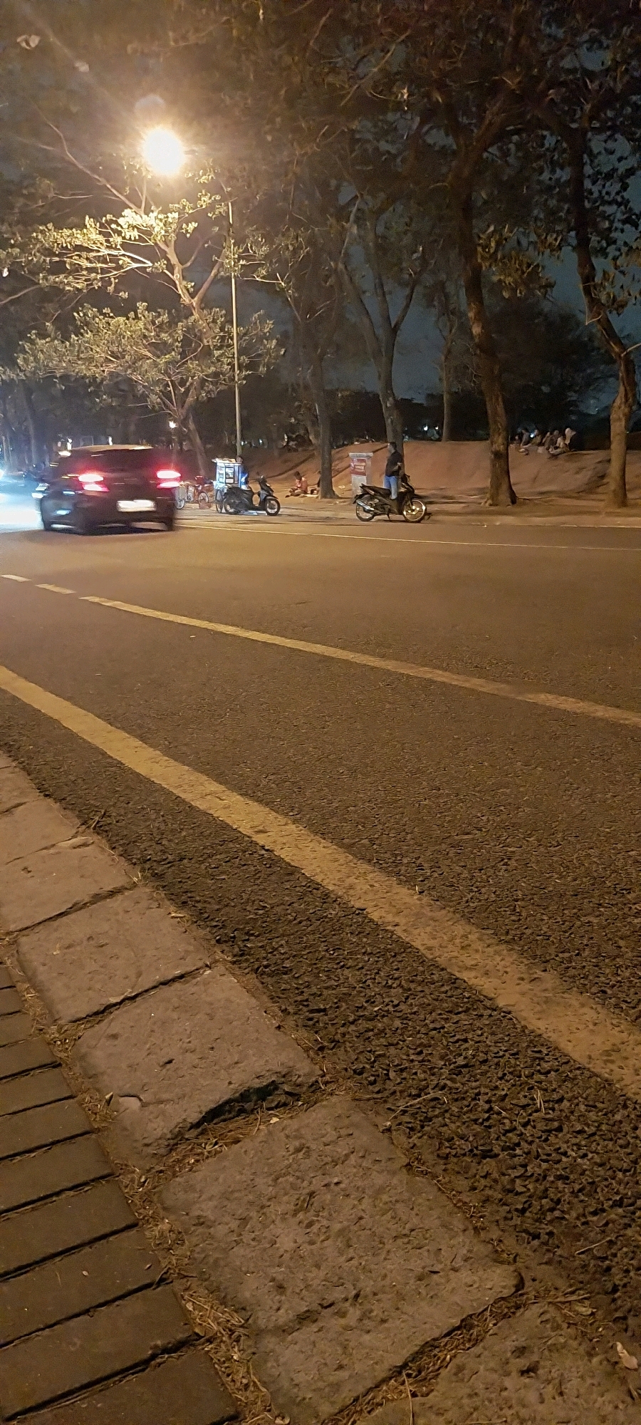 The image captures a nighttime street scene in Kelapa Dua, Indonesia. A dark-colored car, possibly a sedan, is in motion on the asphalt road, its taillights illuminated. The road is marked with two yellow lines. To the left of the road, there is a sidewalk constructed of interlocking bricks and larger, uneven concrete slabs, with dry grass and debris scattered between them. Beyond the road, a pedestrian area is visible, lined with trees and illuminated by a street lamp. Several motorcycles are parked in this area, and some people are gathered or seated on a raised embankment. One motorcycle is parked near a food cart. The overall atmosphere is dimly lit by artificial light sources, with the sky appearing dark and overcast.