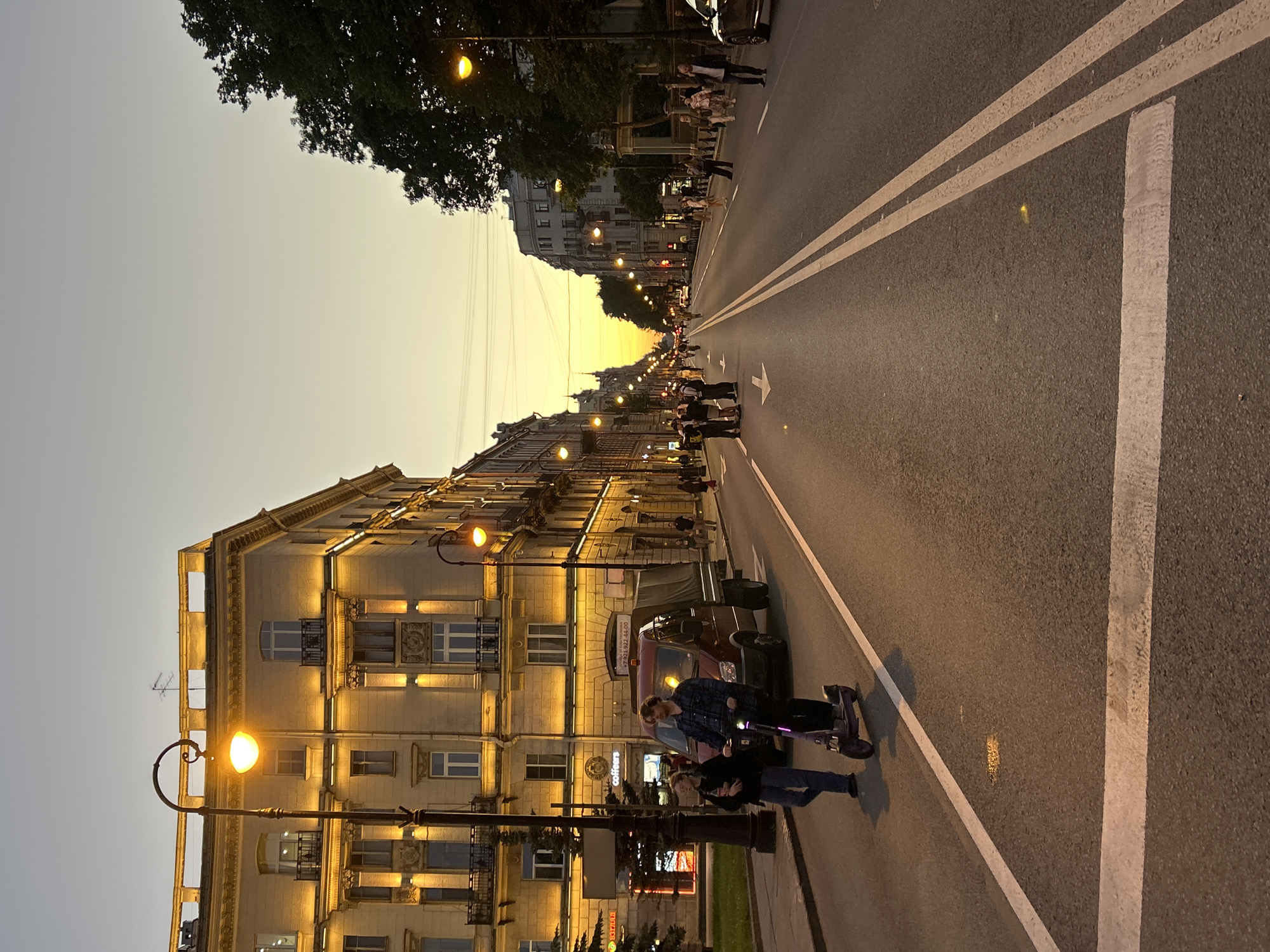 This image depicts a street scene in an urban environment during twilight. The sky is a gradient of pale yellow to gray. On the left side of the street, a multi-story building with ornate architectural details and lit windows is visible. Streetlights with glowing yellow lamps illuminate the sidewalk and street. A dark-colored truck with a canvas cover is parked on the side of the road. In the foreground, two individuals are walking on the sidewalk, one of whom is riding an electric scooter. The street is paved and has white lane markings. People are also seen walking on the sidewalk on the right side of the street and in the distance. Trees line the right side of the street, and other buildings are visible further down the road. The overall atmosphere suggests a mild evening in a city with active pedestrian and vehicle traffic.