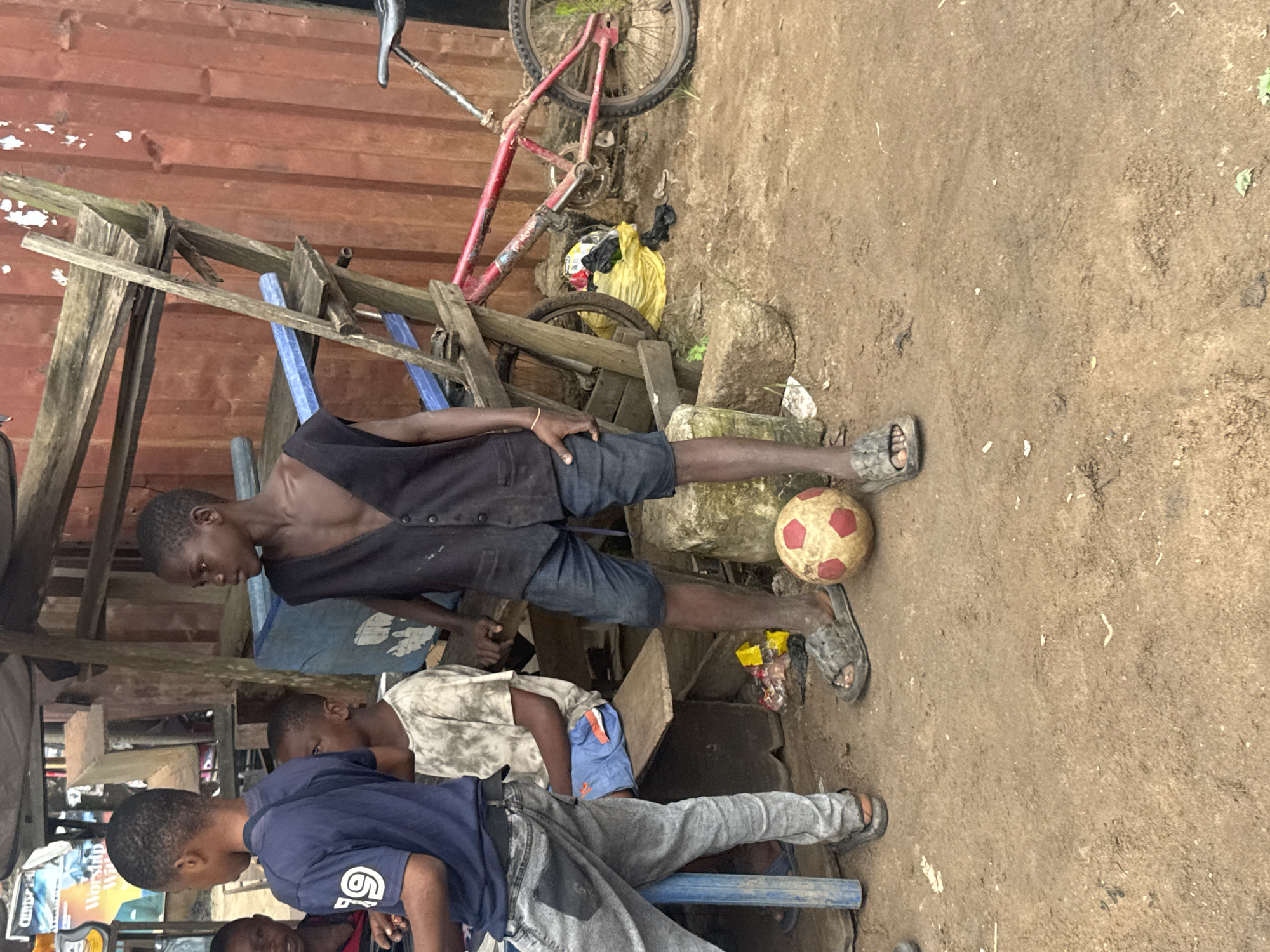 In Ihiagwa, Nigeria, a group of young boys are gathered in a sandy, outdoor area. The central figure, a boy wearing a dark vest and denim shorts, stands prominently with a soccer ball at his feet. Another boy, to his left, is dressed in a blue t-shirt with the number "19" on it and grey jeans. A third boy, seated on a wooden bench, wears a tie-dye t-shirt and blue shorts. The environment is rustic, featuring a makeshift wooden structure with shelves, a red corrugated metal wall in the background, and a red bicycle leaning against the structure. The ground is composed of loose dirt and sand, with some debris, including a yellow plastic bag, scattered around.