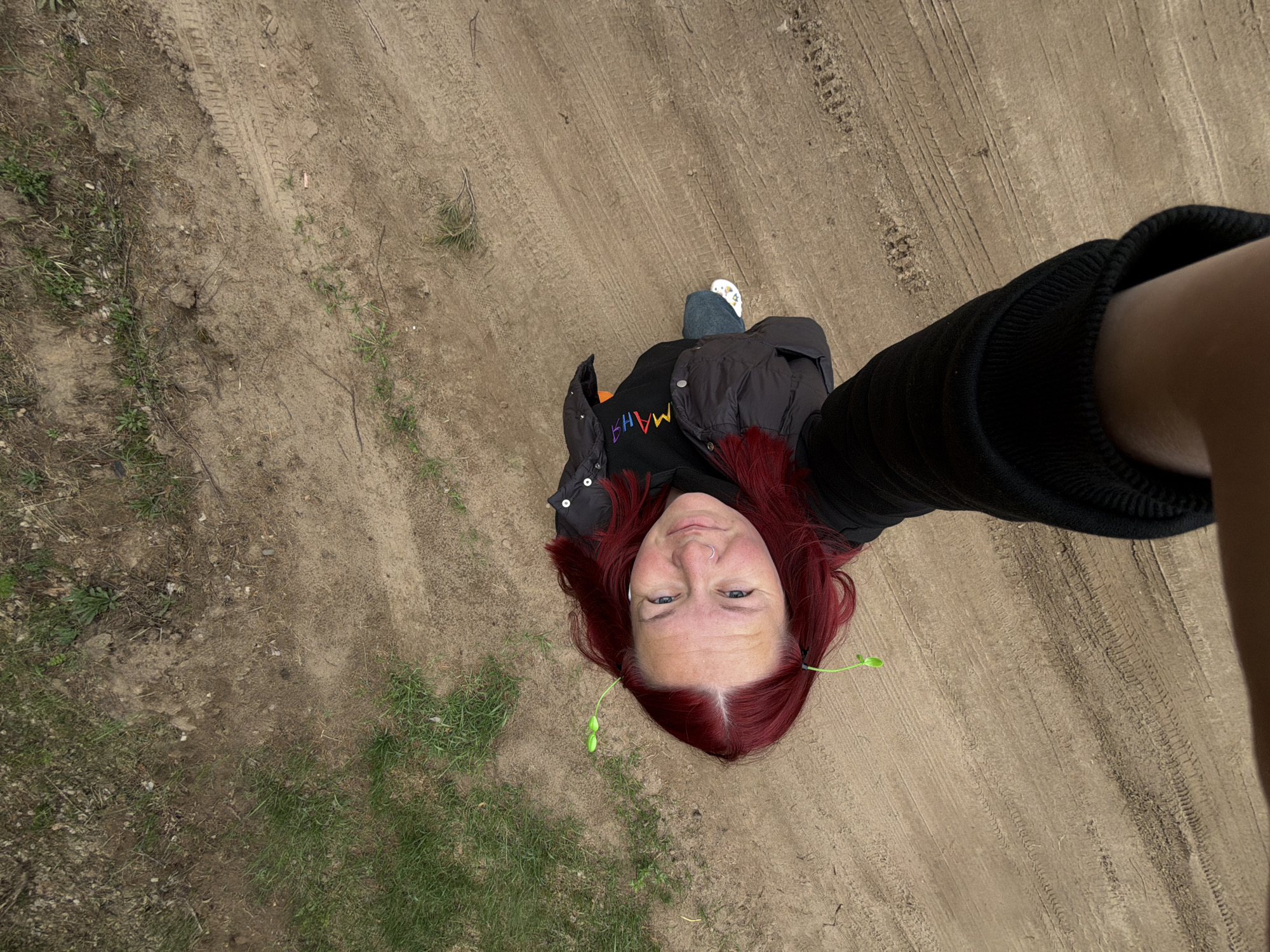 A high-angle, wide-shot photograph depicts a woman in an outdoor setting, possibly in Chaikovsky, Russia. She is standing on a dirt path, interspersed with tire tracks, and patches of grass or vegetation on the edges. The woman has red hair and is wearing a black jacket, a partially visible dark-colored shirt, and blue jeans. Her footwear includes white shoes with designs. She appears to be taking a selfie with an extended arm. She has green sprouts in her hair.