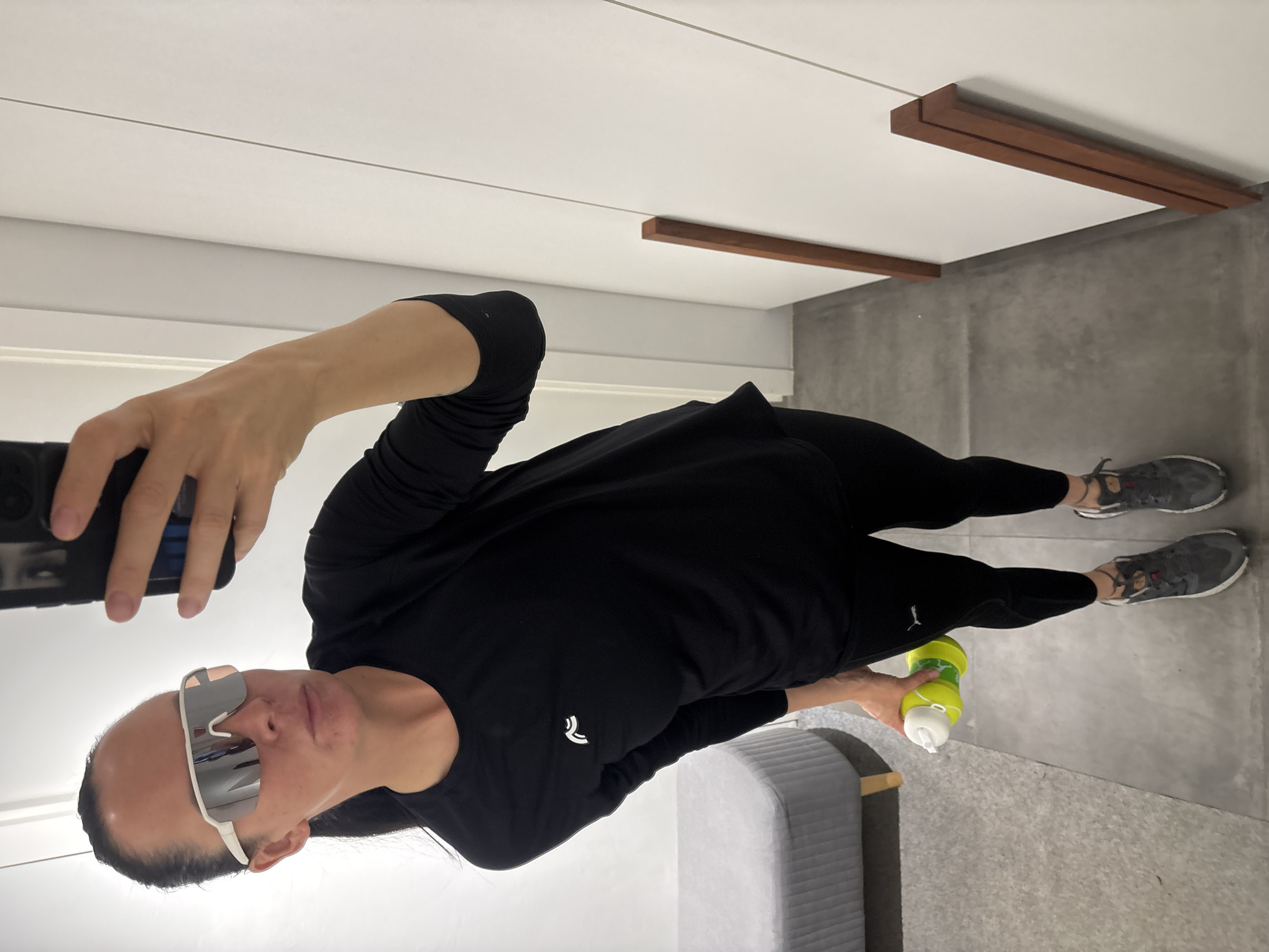 A person wearing black athletic clothing, including leggings and a long-sleeved shirt, and mirrored sunglasses is taking a selfie in what appears to be an indoor space. They are holding a smartphone in one hand and a yellow and white water bottle in the other. The person is standing on a gray concrete floor near a light gray piece of furniture, possibly a couch. Overhead, there is a white ceiling with linear lighting fixtures and two brown wooden accents attached to the ceiling. Without additional evidence, it is not possible to identify the exact location in Chaikovsky, Russia.