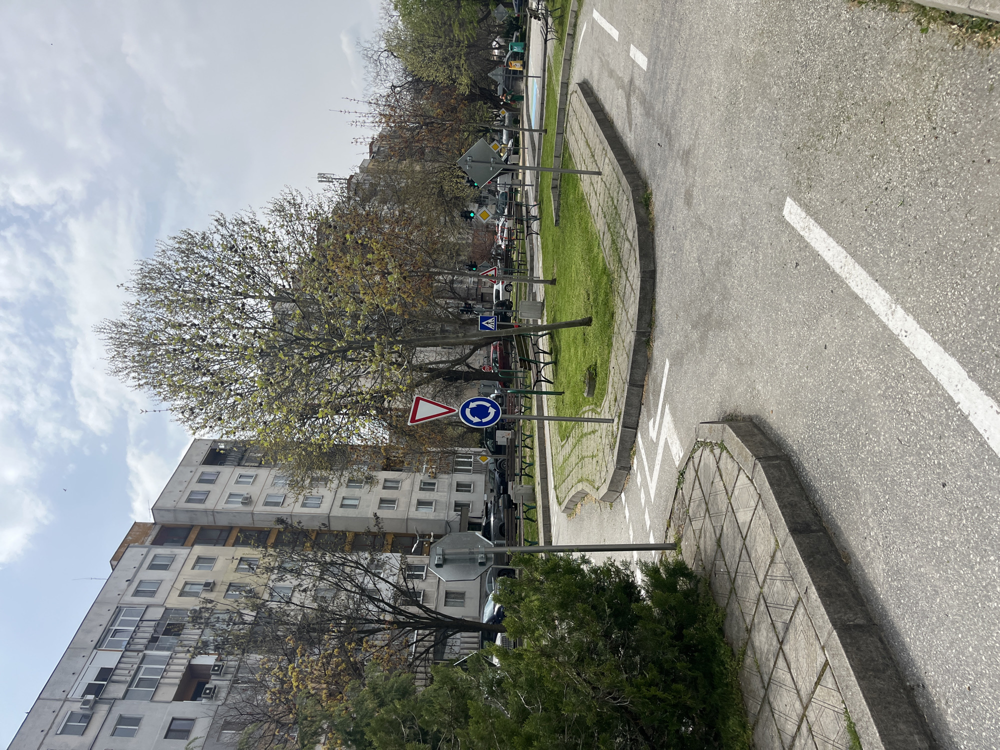 The image depicts a street scene in Plovdiv, Bulgaria, featuring a mock roundabout with traffic signs, including a yield sign and a circular road sign. A multi-story residential building stands to the left of the roundabout. The road is marked with white lines. Vegetation, including grass and trees, surrounds the roundabout. Cars are parked behind the roundabout. The sky is cloudy.